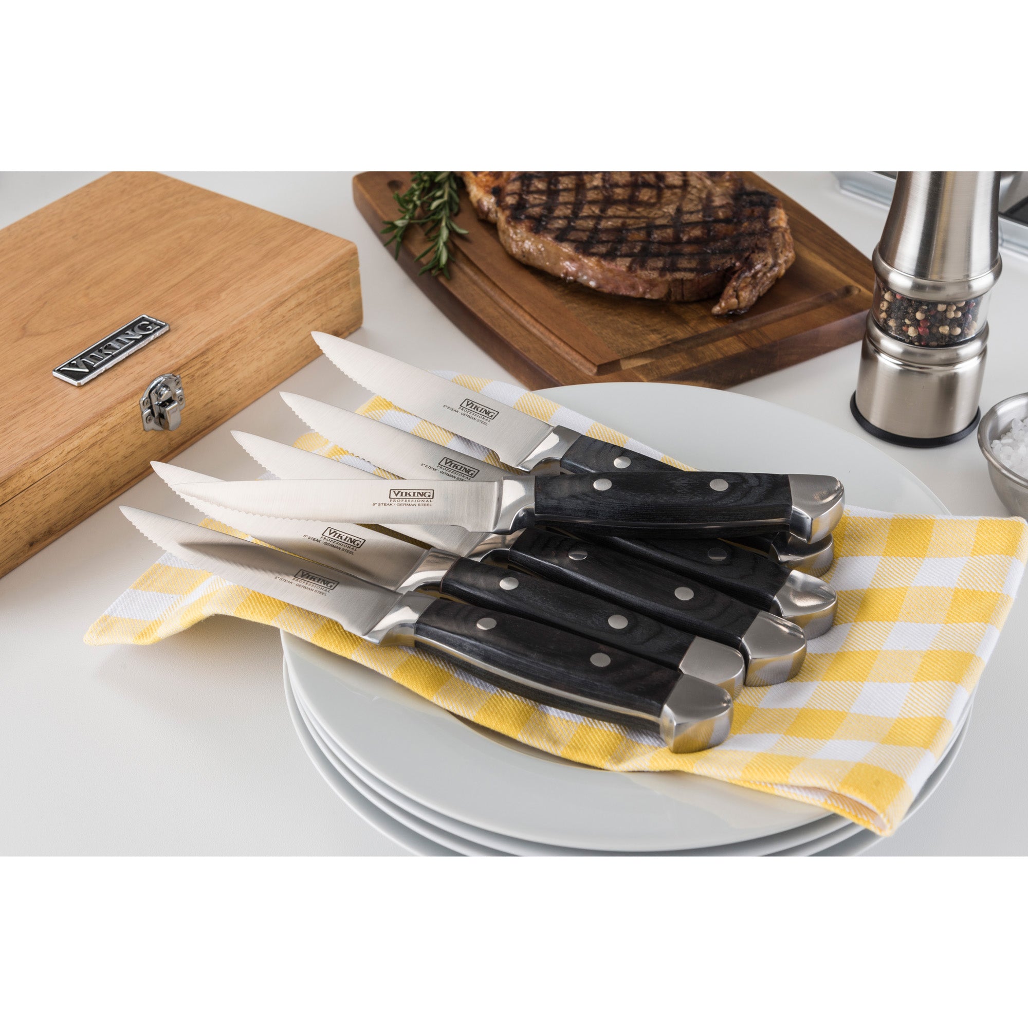 The Viking Steakhouse Pakka Wood 6-Piece Steak Knife Set by Viking Culinary Products features corrosion-resistant black pakkawood handles and comes in a gift box, shown here arranged beside a grilled steak and pepper grinder.