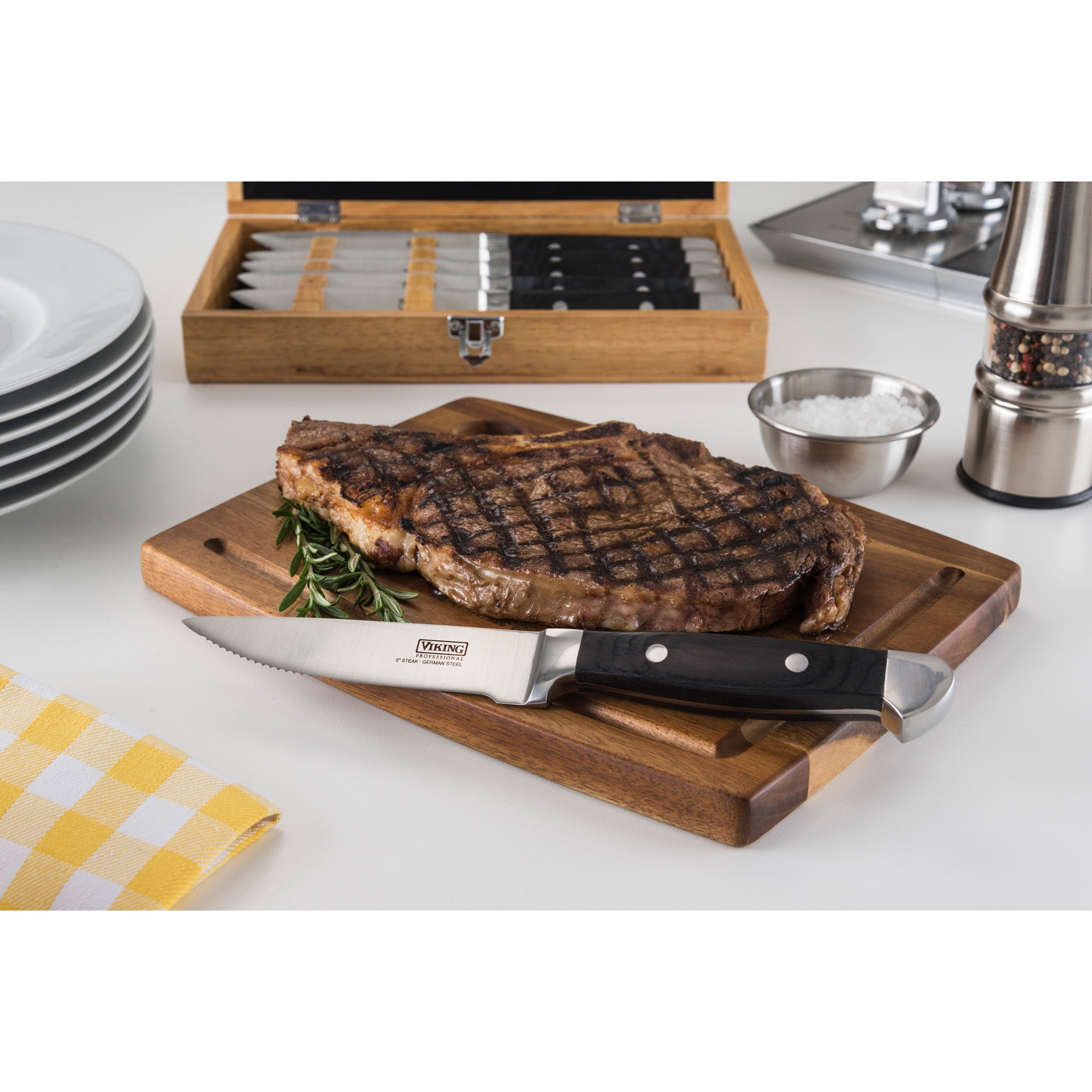 A grilled steak rests on a wooden board beside a Viking Steakhouse Pakka Wood 6-Piece Steak Knife from Viking Culinary Products. In the background are stacked plates, the corrosion-resistant gift box, salt bowl, pepper grinder, and a yellow checkered napkin.