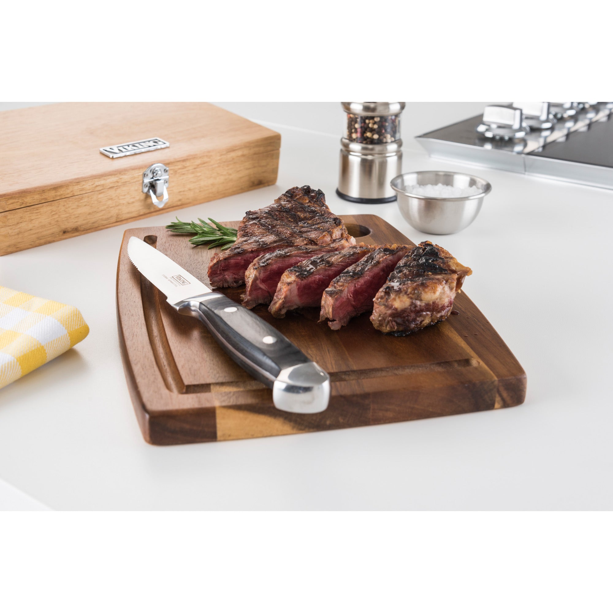 Sliced grilled steak on a wooden cutting board with a Viking Culinary Products Viking Steakhouse Pakka Wood 6-Piece Steak Knife Set (Black) in its gift box, rosemary, salt bowl, pepper grinder, and a yellow checkered napkin on a white countertop.