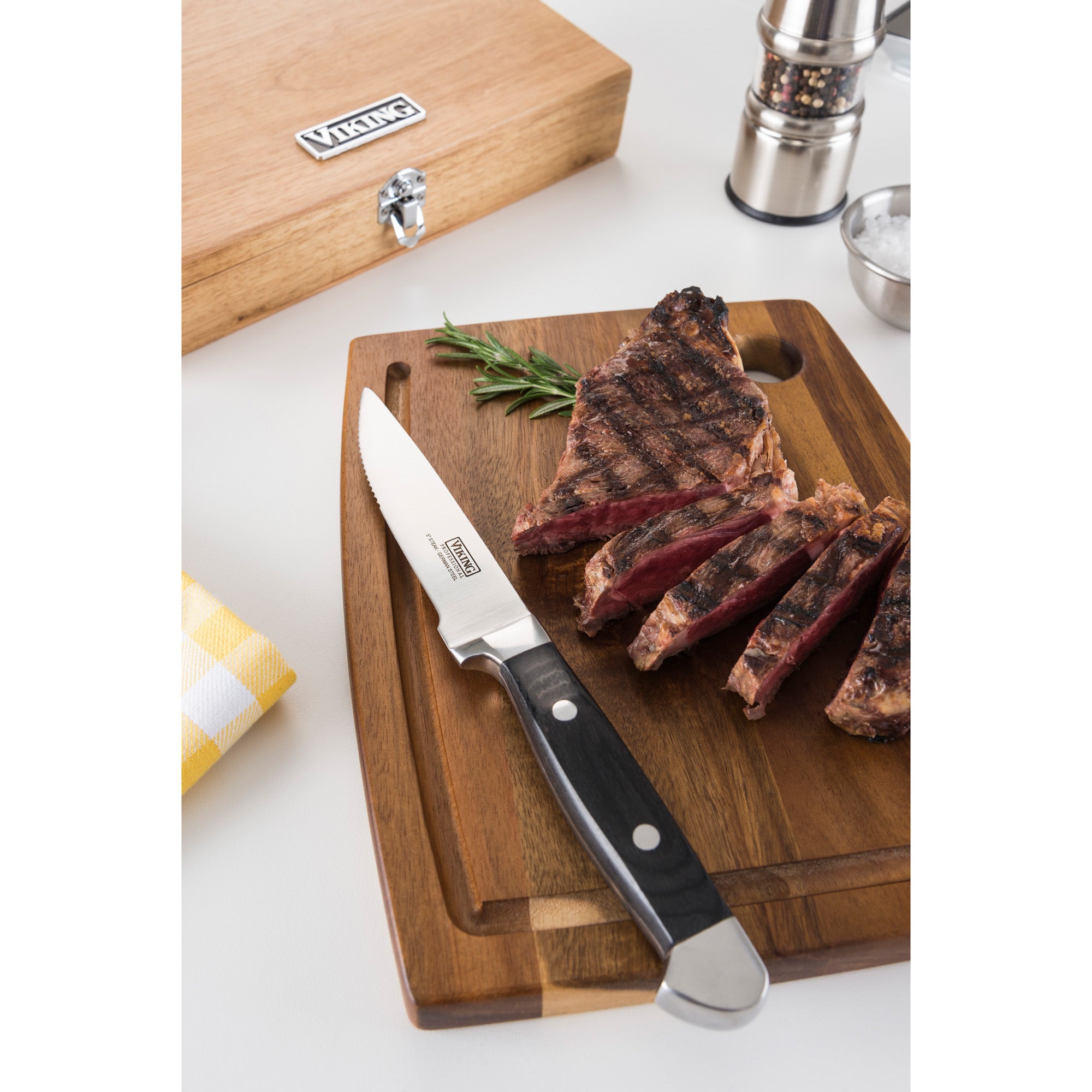 A grilled steak, partially sliced with Viking Steakhouse Pakka Wood 6-Piece Steak Knife Set by Viking Culinary Products, sits on a wooden board. Nearby are a branded black gift box, salt, pepper grinder, rosemary sprig, and yellow-checked napkin.