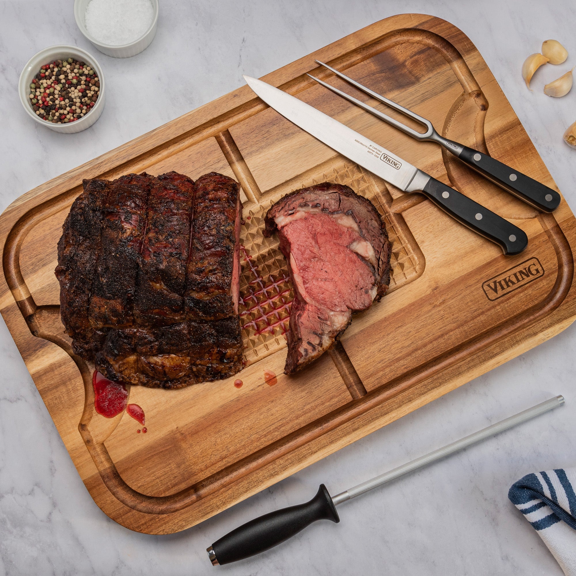 A juicy roast beef sits on the Viking Acacia Wood Cutting Board with the 3-Piece Stainless Steel Carving Set by Viking Culinary Products, surrounded by salt, peppercorns, garlic cloves, and a striped towel on a marble surface.