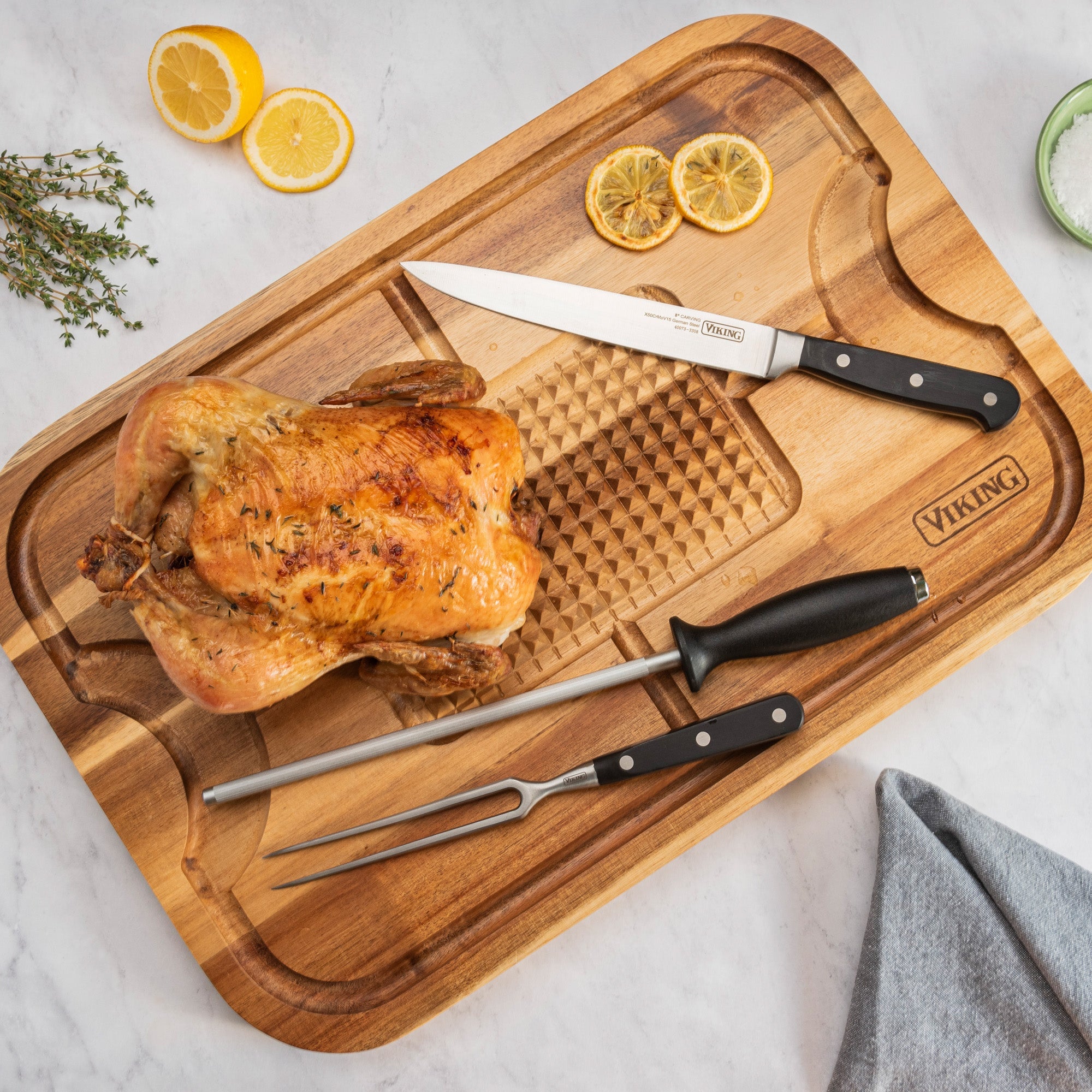 A roasted chicken sits on the Viking Acacia Wood Cutting Board with 3-Piece Stainless Steel Carving Set by Viking Culinary Products, surrounded by lemon slices, herbs, and a bowl of salt on a marble surface.
