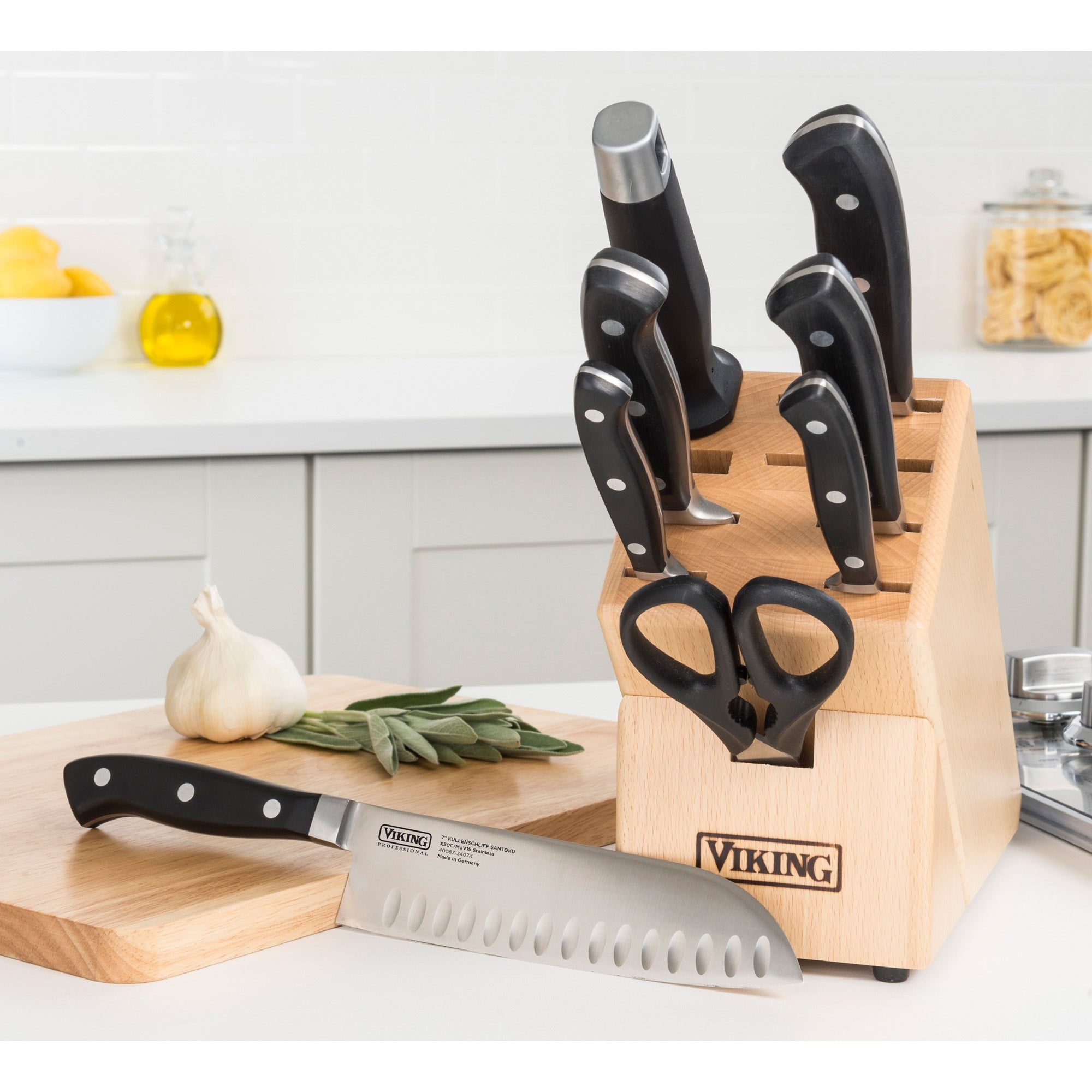 The Viking Culinary Products Professional 7-Piece Cutlery Set features German steel, full-tang knives in a wooden block, plus kitchen scissors and a chef knife on a cutting board with garlic and sage in a modern kitchen setting.