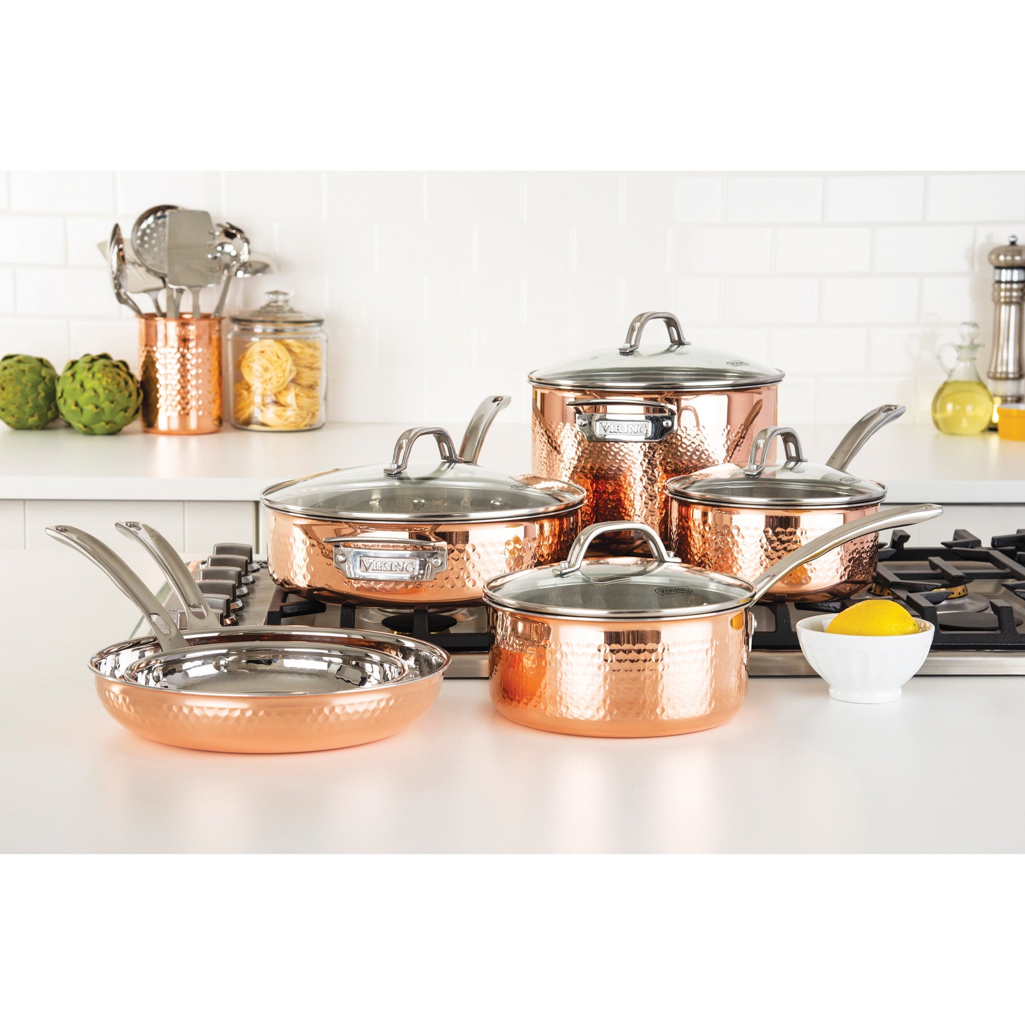 The Viking 3-Ply Hammered Copper Clad 10-Piece Cookware Set with Glass Lids by Viking Culinary Products sits on a white kitchen counter with a lemon, artichokes, utensils, and jars, against a clean white tile backsplash.