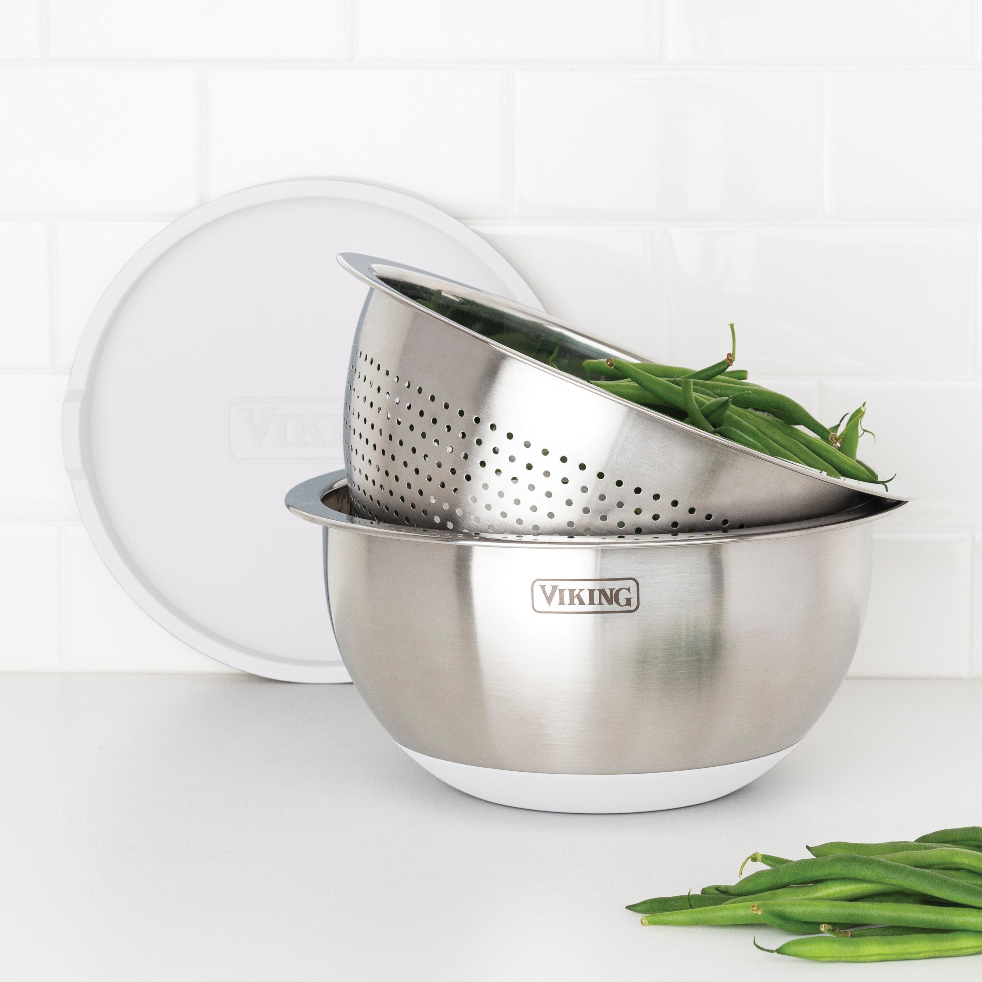 The Viking Culinary Products 9-Piece Stainless Steel Mixing Bowl Set with Strainer in Frost White is displayed on a white counter with its lid, featuring green beans in the strainer nested inside a mixing bowl against a white tiled backsplash.