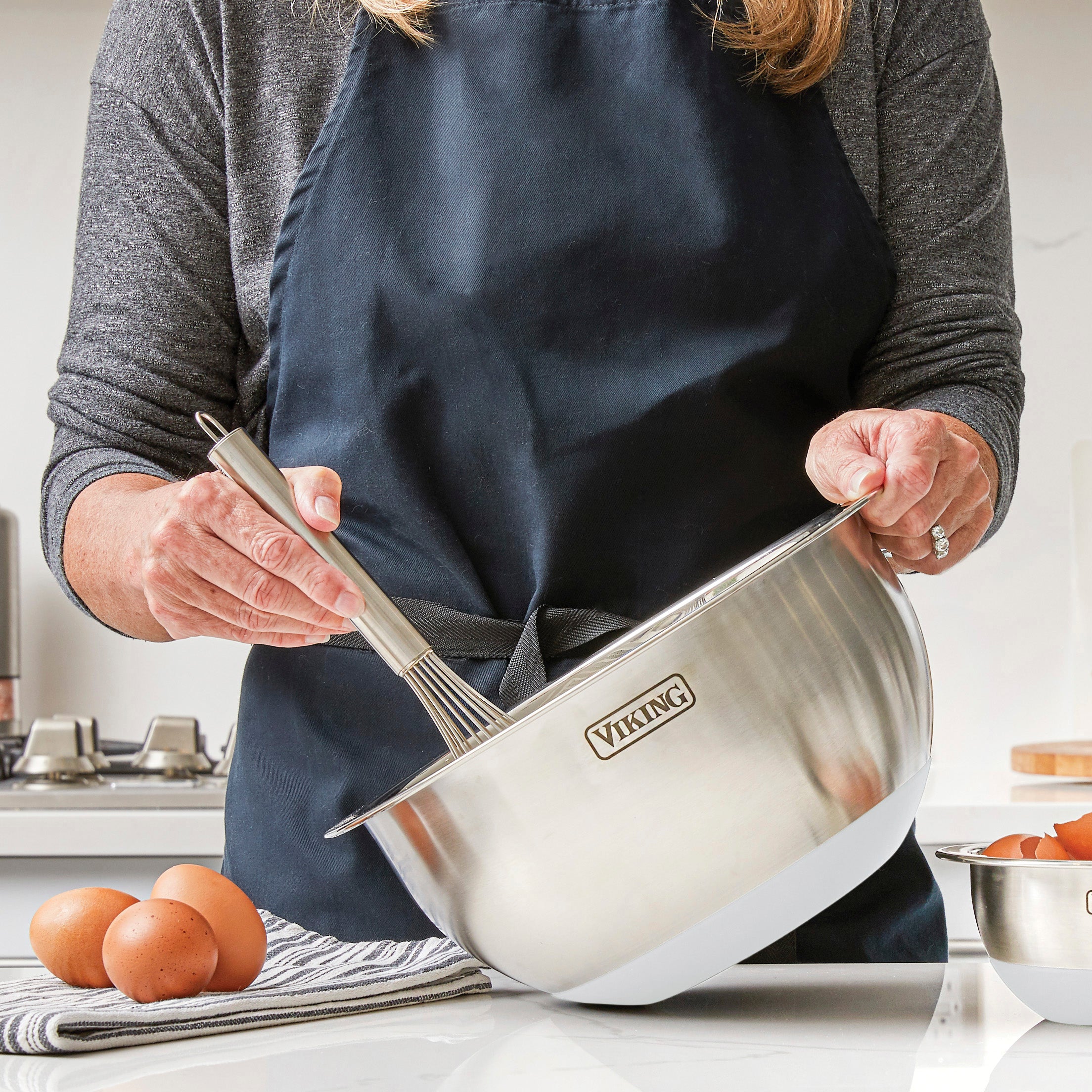 A person in a dark apron whisks ingredients in a Viking Culinary Products 9-Piece Stainless Steel Mixing Bowl with Strainer, Frost White, while eggs and nesting bowls sit on the kitchen counter nearby.
