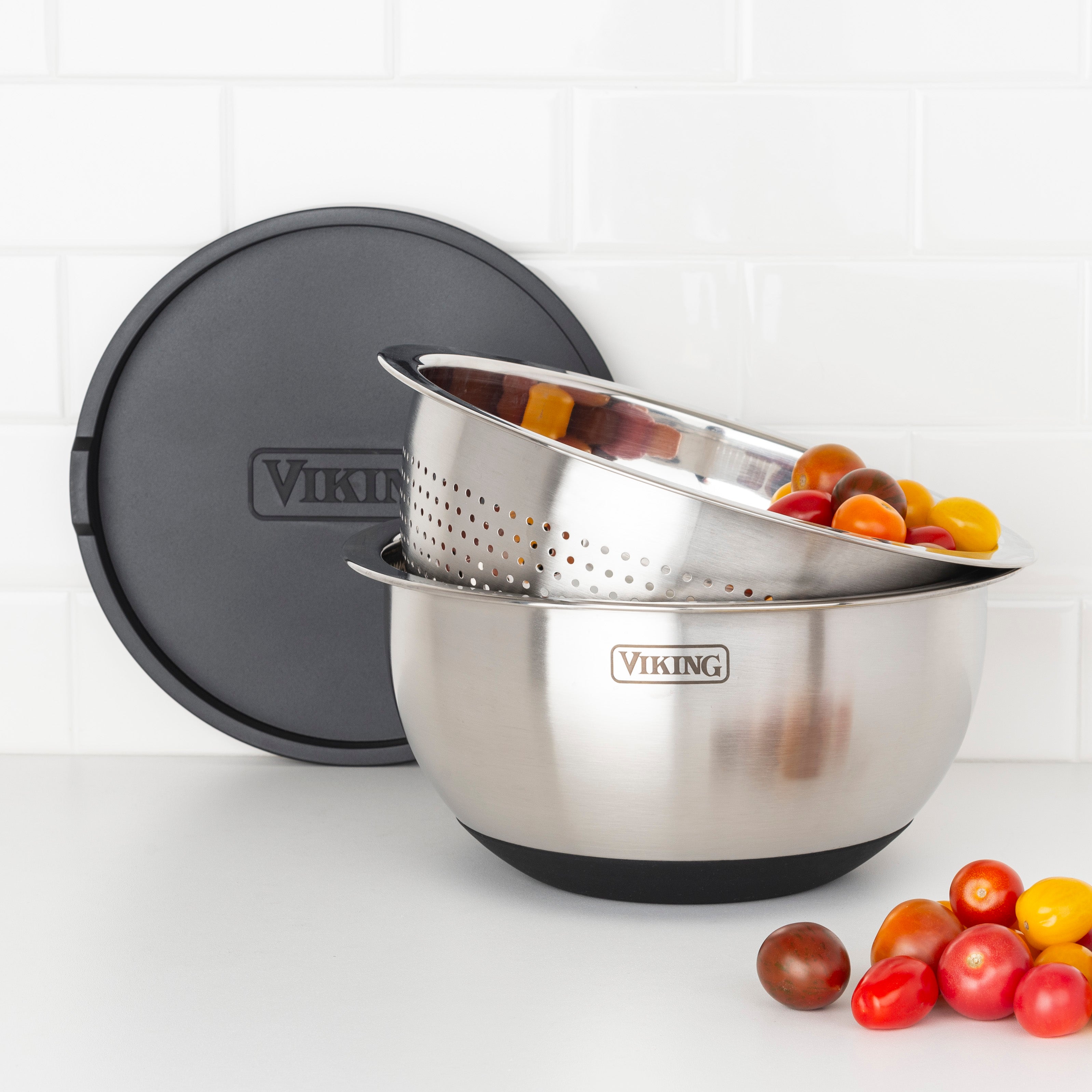 The Viking Culinary Products 9-Piece Stainless Steel Mixing Bowl Set with Strainer in black features airtight lids and silicone bases, shown with cherry tomatoes on a white countertop against a tiled wall.