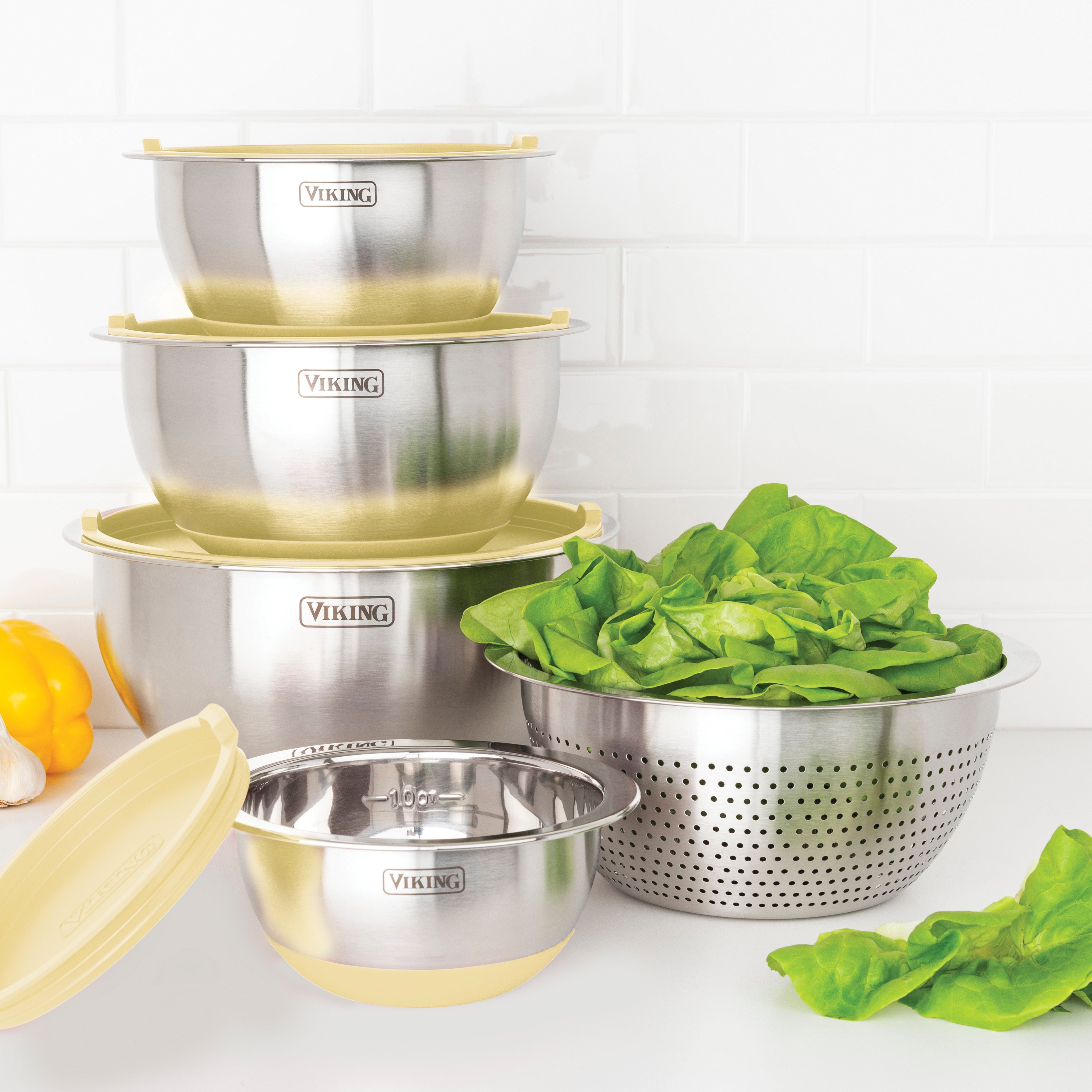 The Viking Culinary Products 9-Piece Stainless Steel Mixing Bowl Set with Strainer in Buttermilk is stacked beside a colander filled with fresh green lettuce, all on a white countertop with a tiled backsplash.