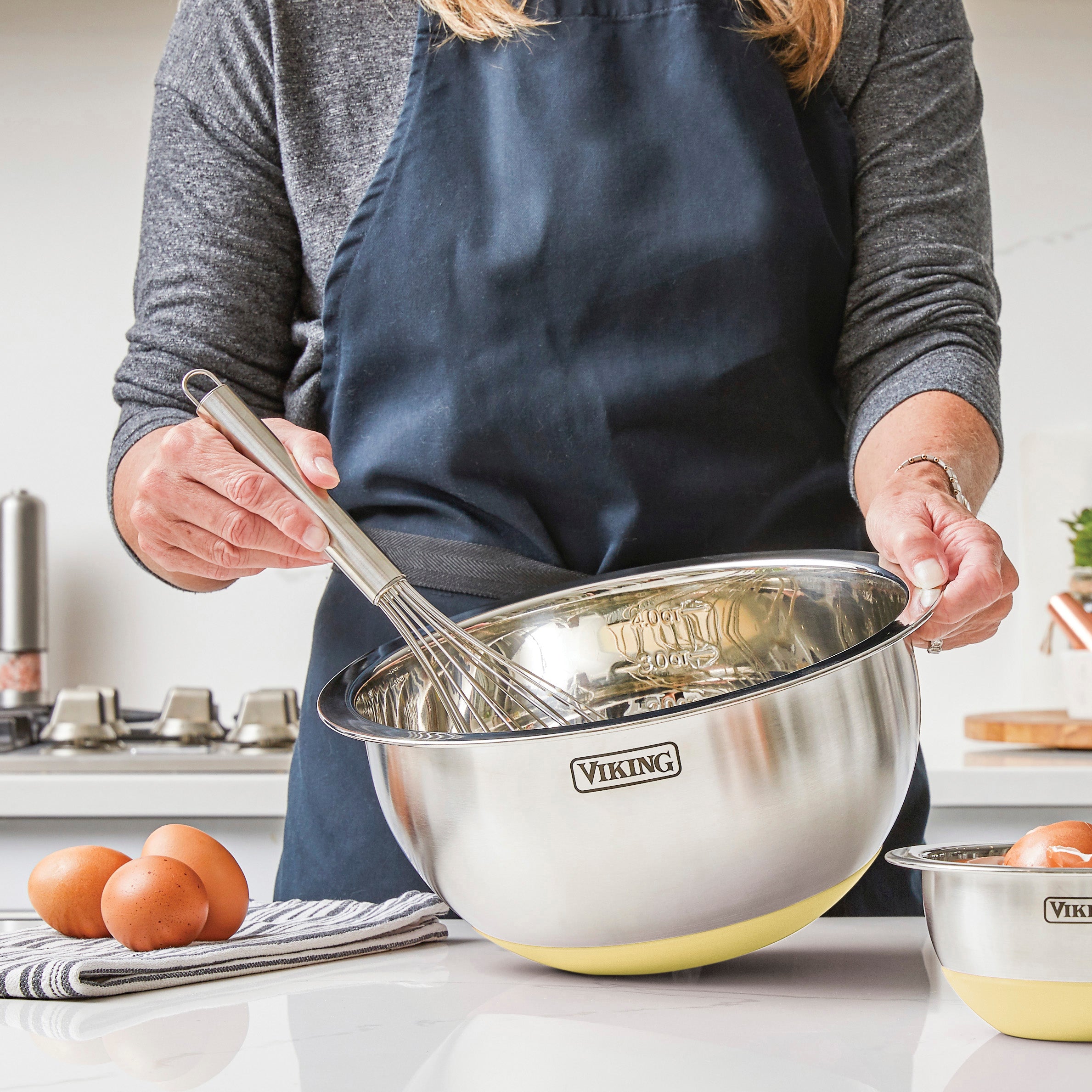 A person in a dark apron is whisking ingredients in a large Viking Culinary Products stainless steel bowl, part of the 9-Piece Mixing Bowl Set with Strainer (Buttermilk), on a kitchen counter with eggs and a striped towel nearby.