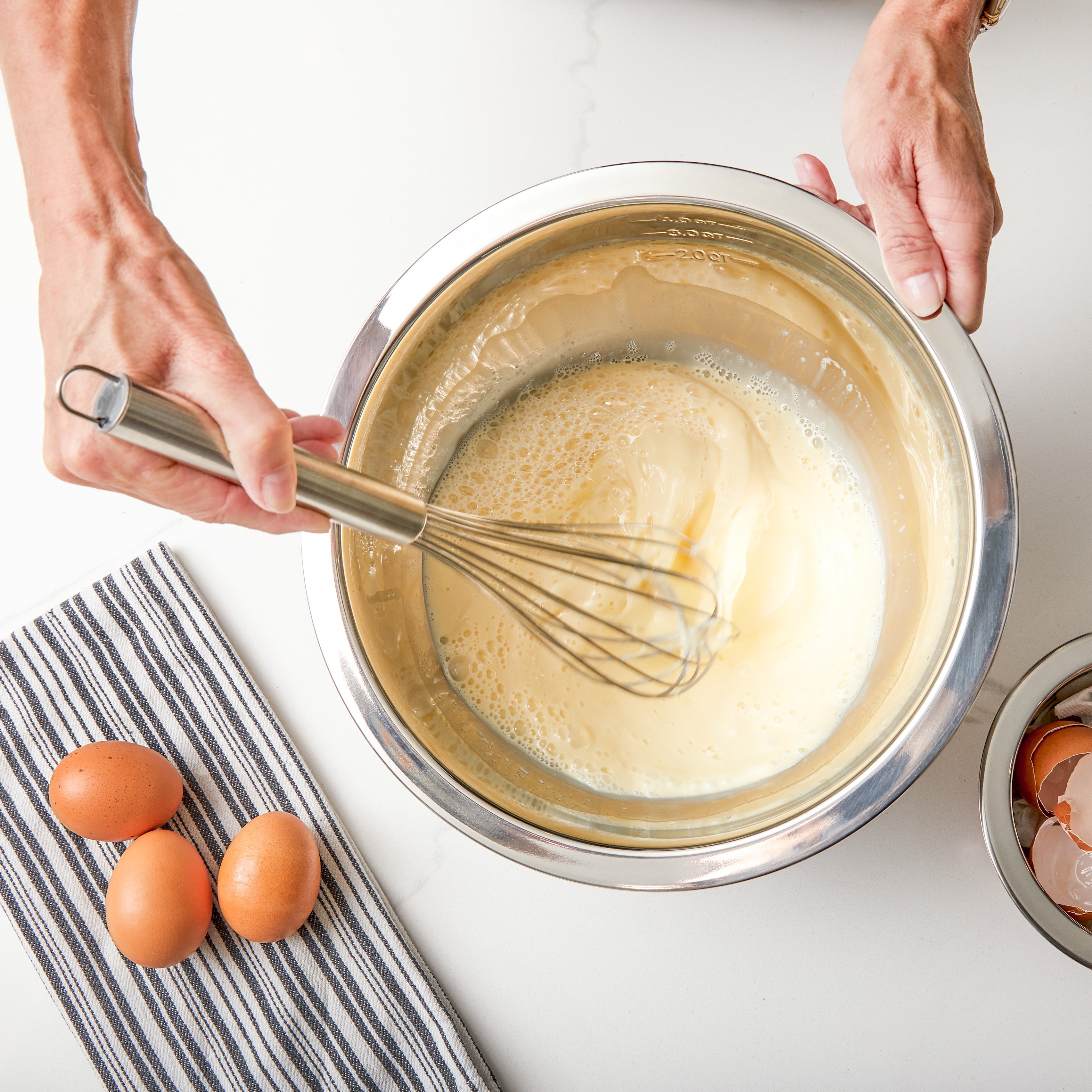 A person whisks a pale yellow mixture in a Viking 9-Piece Stainless Steel Mixing Bowl by Viking Culinary Products, with three brown eggs, a striped towel, and a bowl of eggshells nearbyâ€”ideal for compact kitchens and featuring airtight lids.