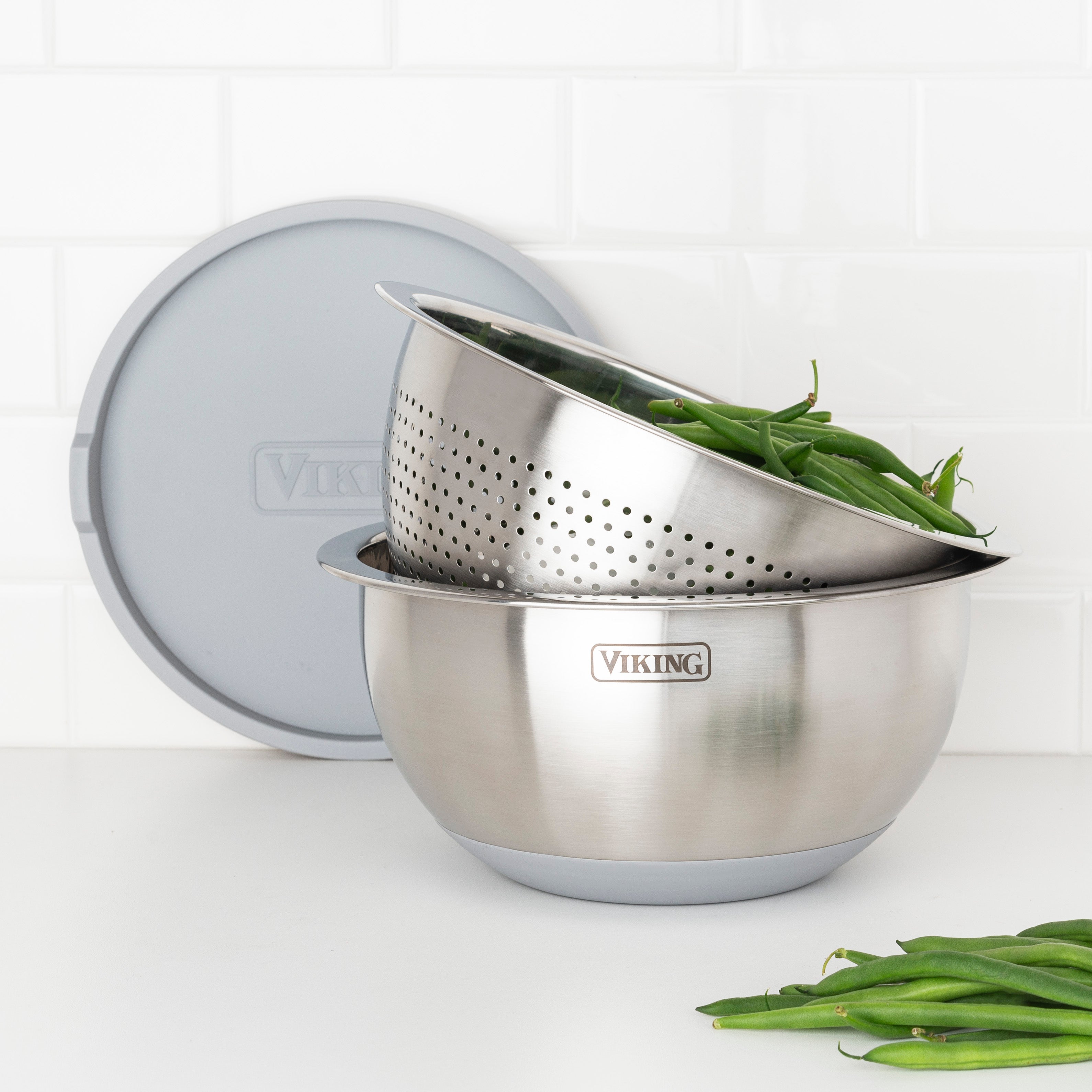 A Viking Culinary Products 9-Piece Stainless Steel Mixing Bowl Set with Strainer in gray sits on a white counter, holding green beans in the colander. These stylish bowls offer compact storage and complement a white tile backsplash.
