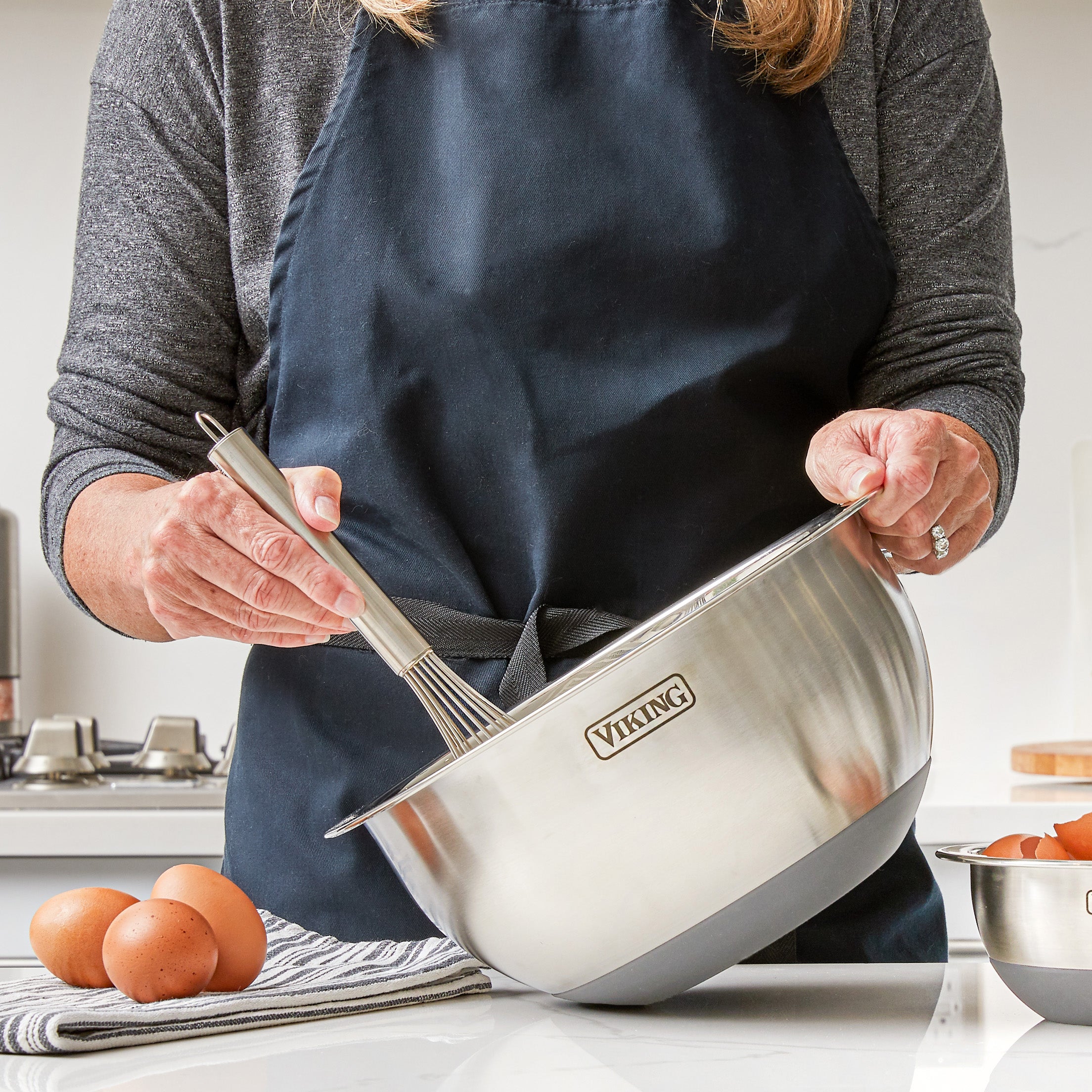 A person in a dark apron is whisking ingredients in a Viking Culinary Products 9-Piece Stainless Steel Mixing Bowl with Strainer, Gray, on a kitchen counter, with eggs and a striped towel nearby.