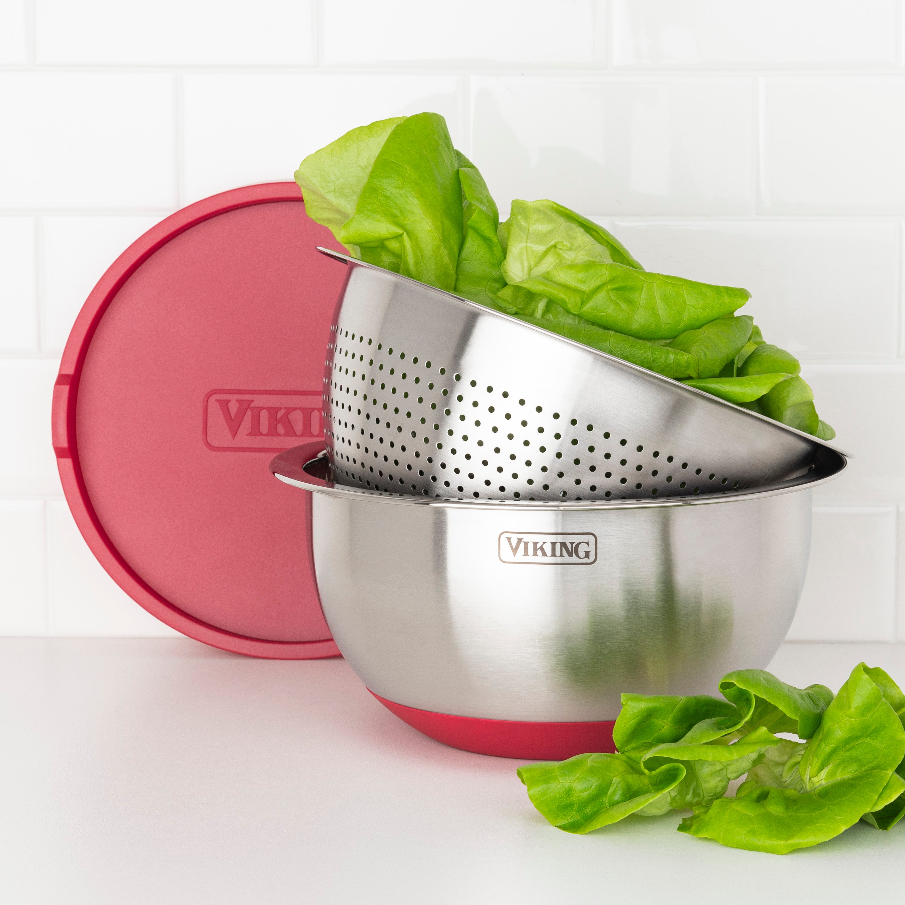 The Viking Culinary Products 9-Piece Stainless Steel Mixing Bowl Set with Strainer, Red, is shown stacked for compact storage with lettuce inside. A red airtight lid leans against the bowls, and lettuce leaves are on the white countertop.