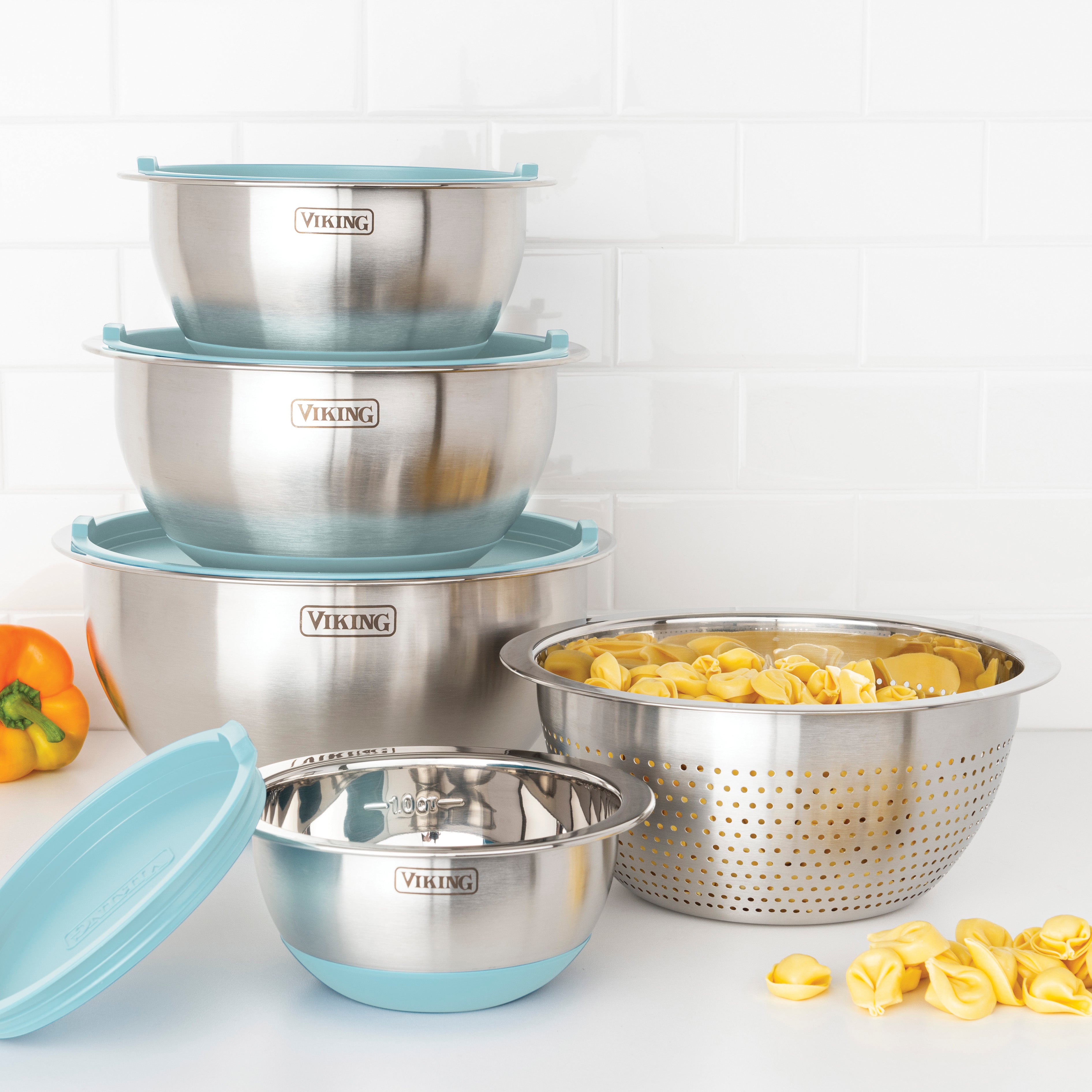 The Viking 9-Piece Stainless Steel Mixing Bowl Set with Strainer in Splash Blue by Viking Culinary Products is stacked on a white kitchen counter, with yellow tortellini and a yellow bell pepper placed nearby.