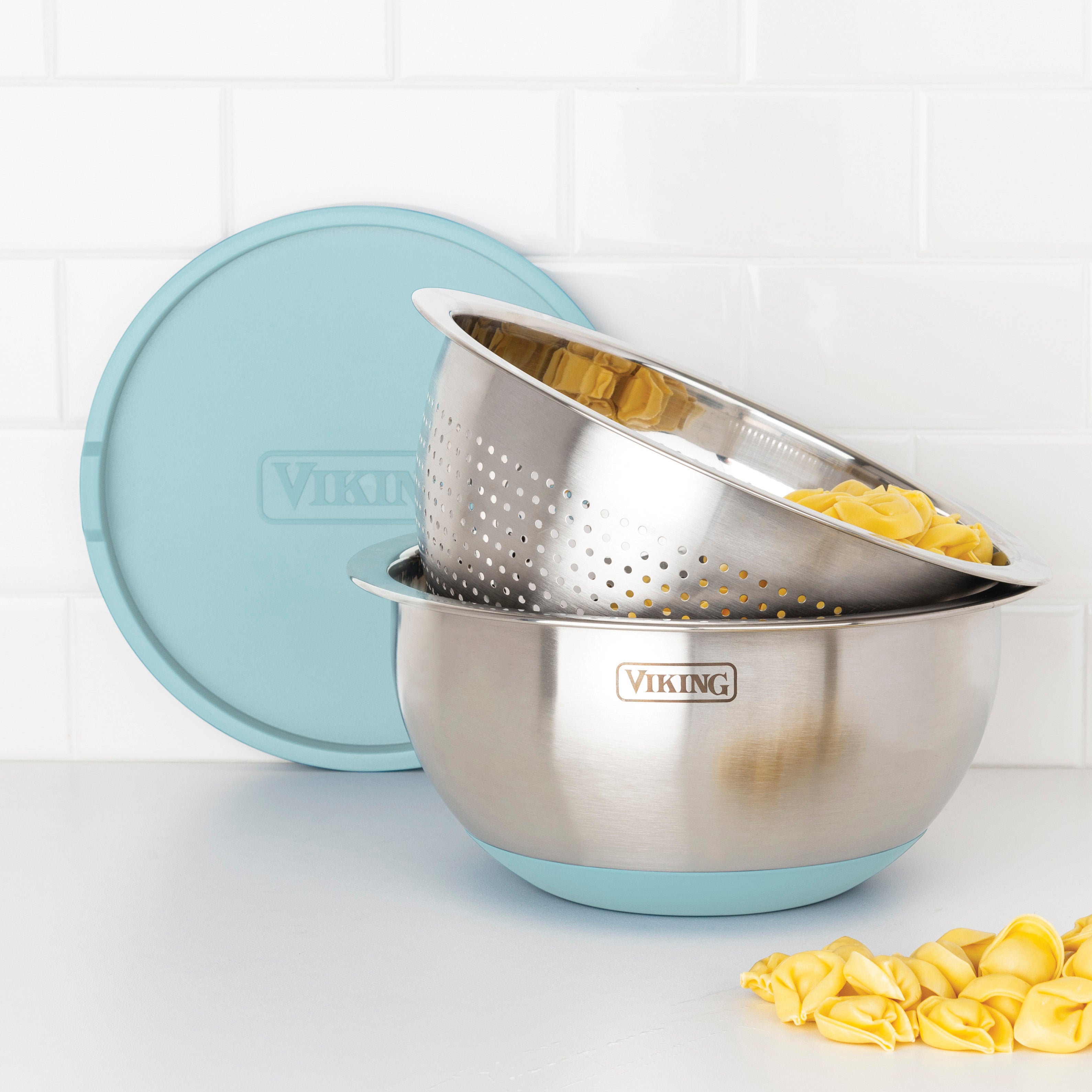 The Viking 9-Piece Stainless Steel Mixing Bowl Set with Strainer in Splash Blue by Viking Culinary Products is stacked on the kitchen counter, with the top colander full of pasta and a few pasta pieces beside the matching lidded bowls.
