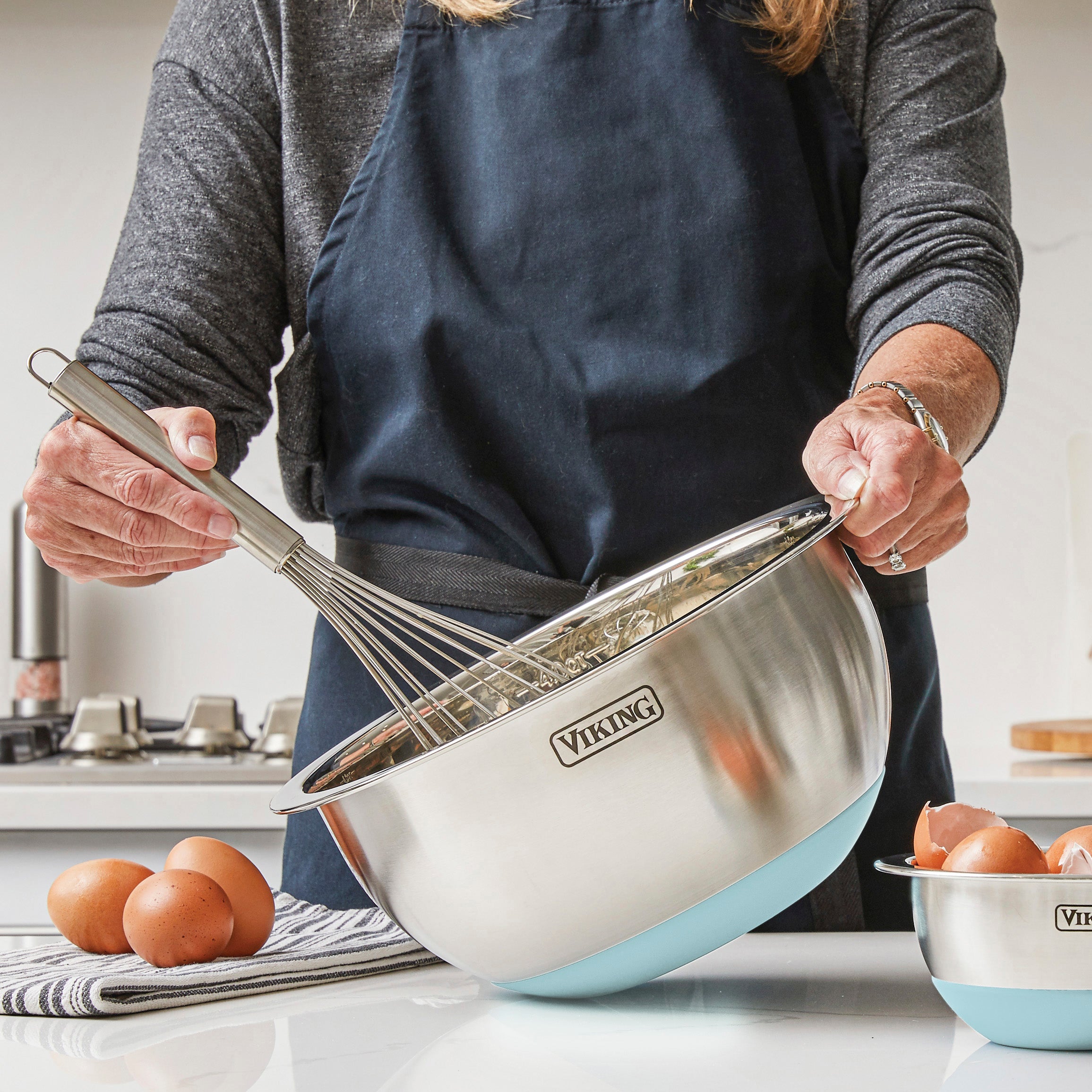 A person wearing a dark apron whisks ingredients in a large bowl from the Viking Culinary Products 9-Piece Stainless Steel Mixing Bowl Set with Strainer, Splash Blue, on a kitchen counter with eggs and other bowls neatly arranged nearby.