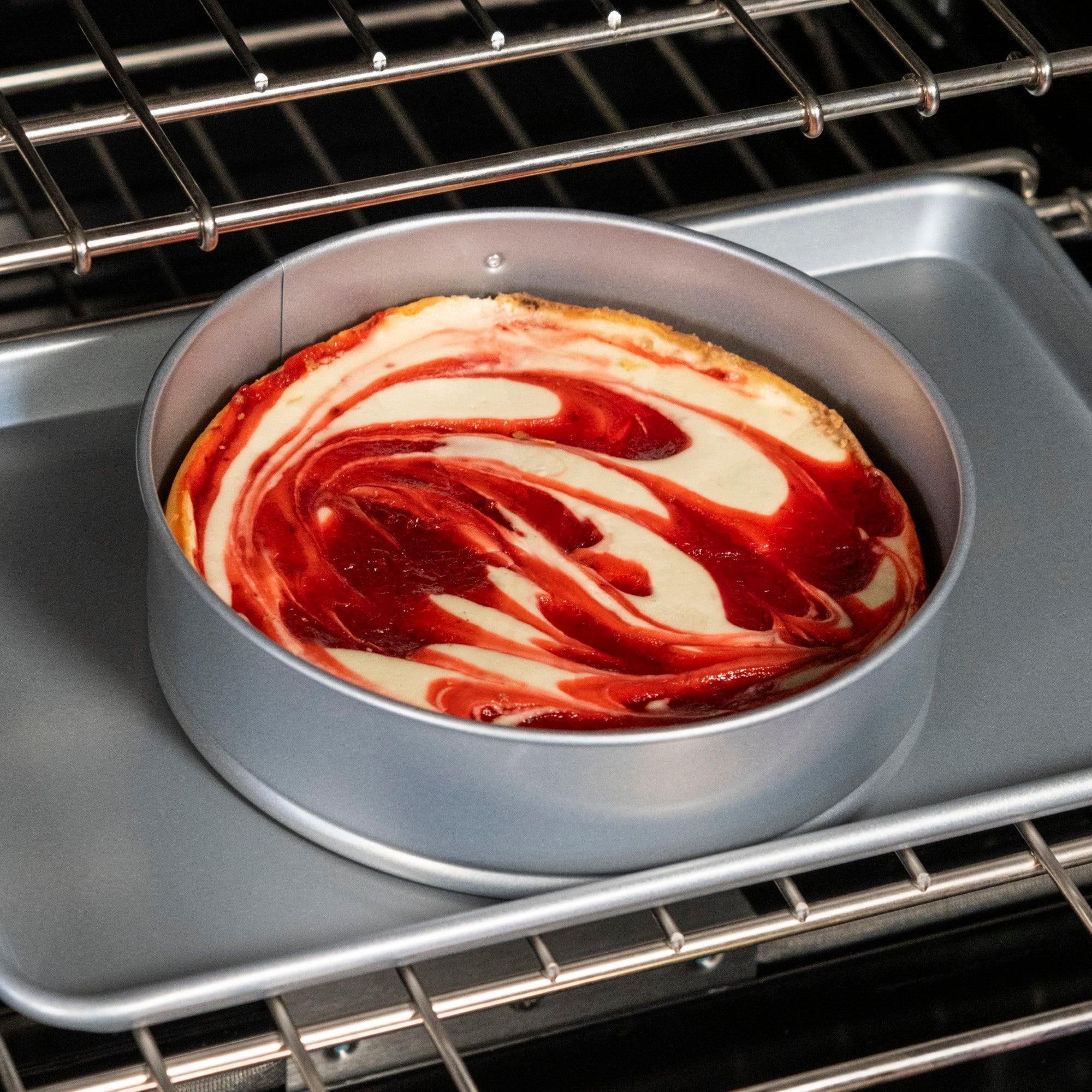 A round cheesecake with red and white swirl topping bakes in a Viking Culinary Products Viking Aluminized Steel Nonstick 9-inch Springform Pan on a baking sheet inside an oven.