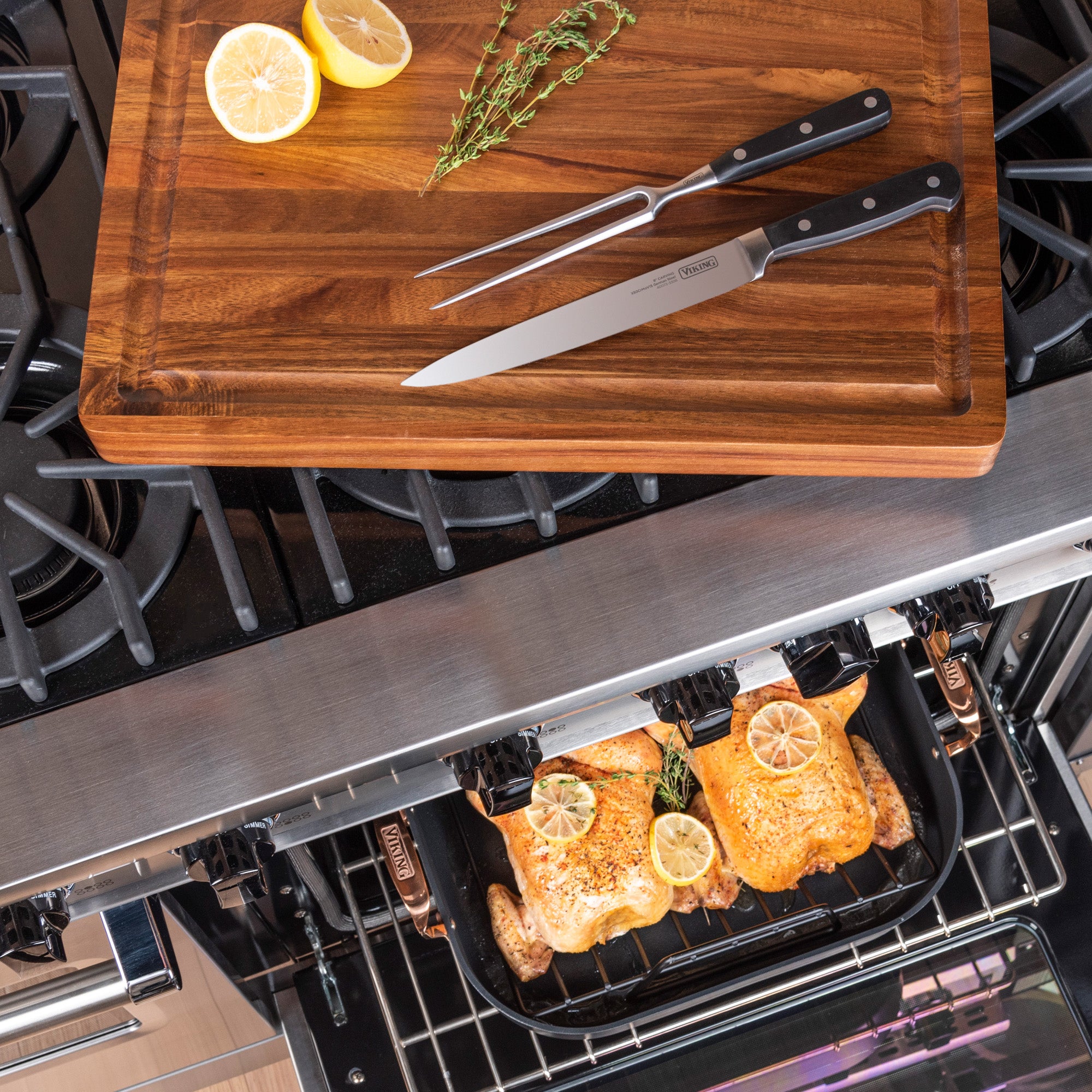 A Viking Hard Anodized Roaster with Copper Handles from Viking Culinary Products holds two roasted chickens topped with lemon and herbs, monitored with the Bluetooth Thermometer, while a carving set and board sit above the oven.