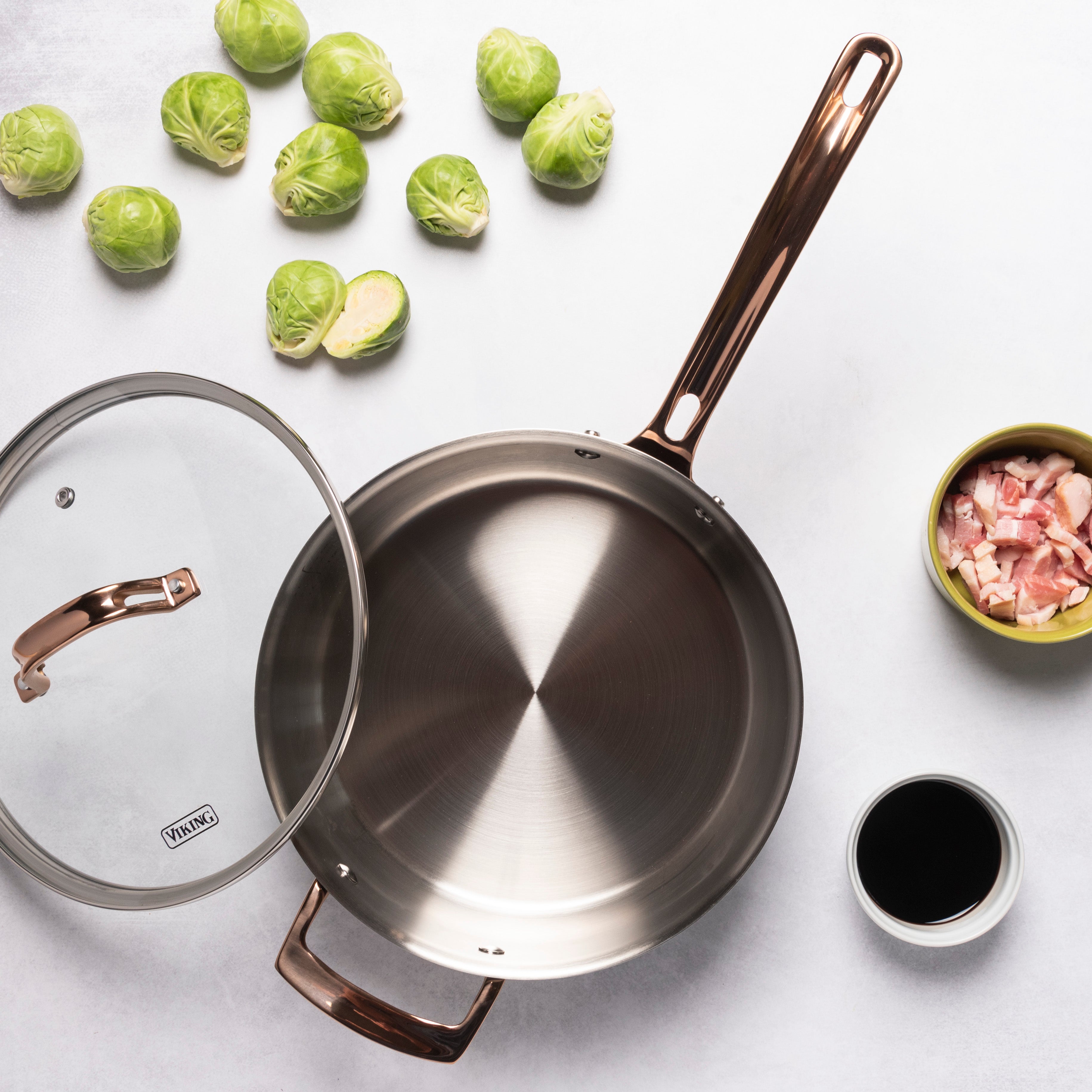 A Viking Culinary Products 3-Ply Black and Copper 4 Quart Sauté Pan with Glass Lid is displayed on a light surface, surrounded by Brussels sprouts, chopped bacon, and a bowl of dark sauce. Induction compatible for versatile stovetop use.