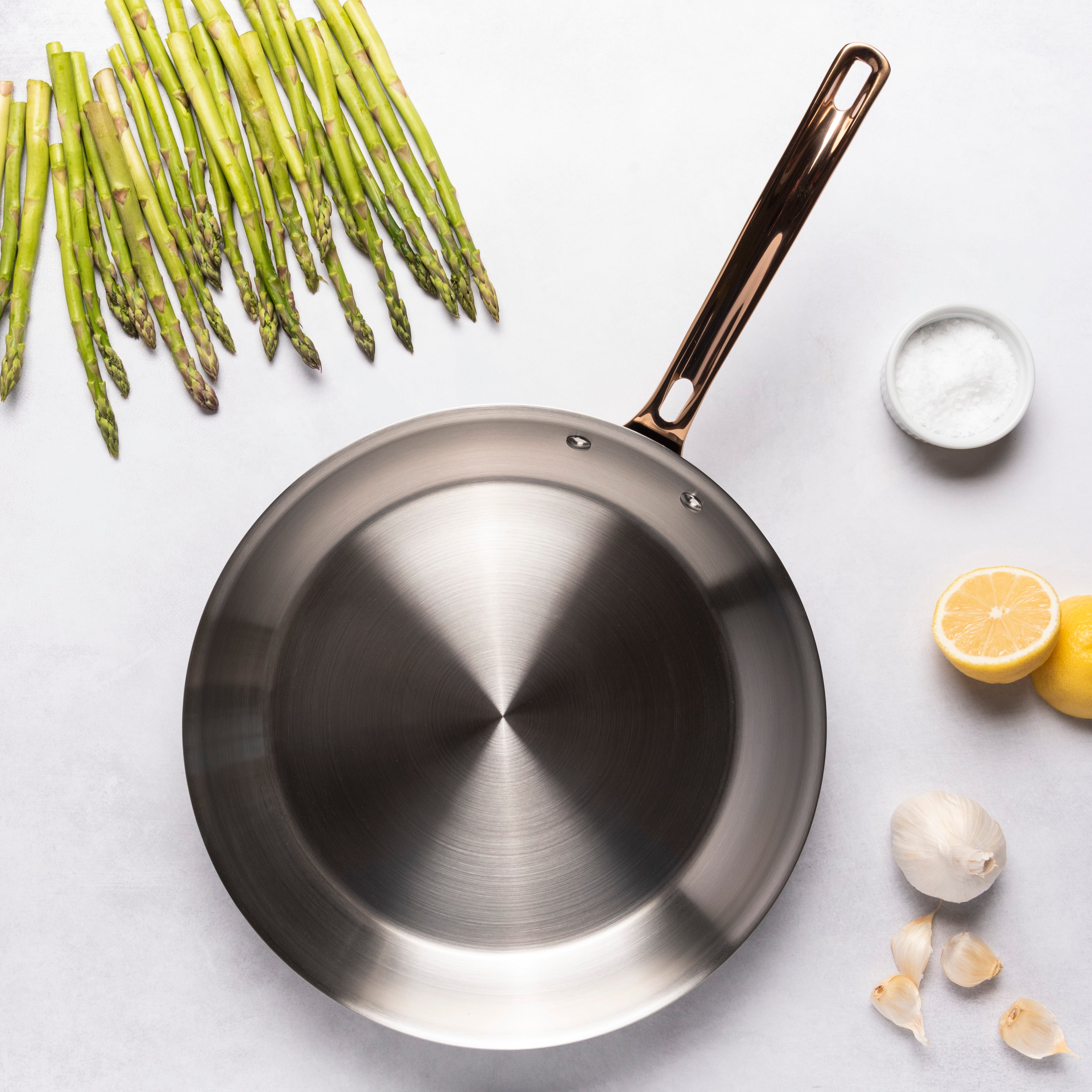 The Viking 3-Ply Black and Copper 12 Inch Fry Pan by Viking Culinary Products, featuring a copper handle, is surrounded by asparagus, a bowl of salt, lemon halves, and garlic cloves on a white surface for optimal heat distribution.