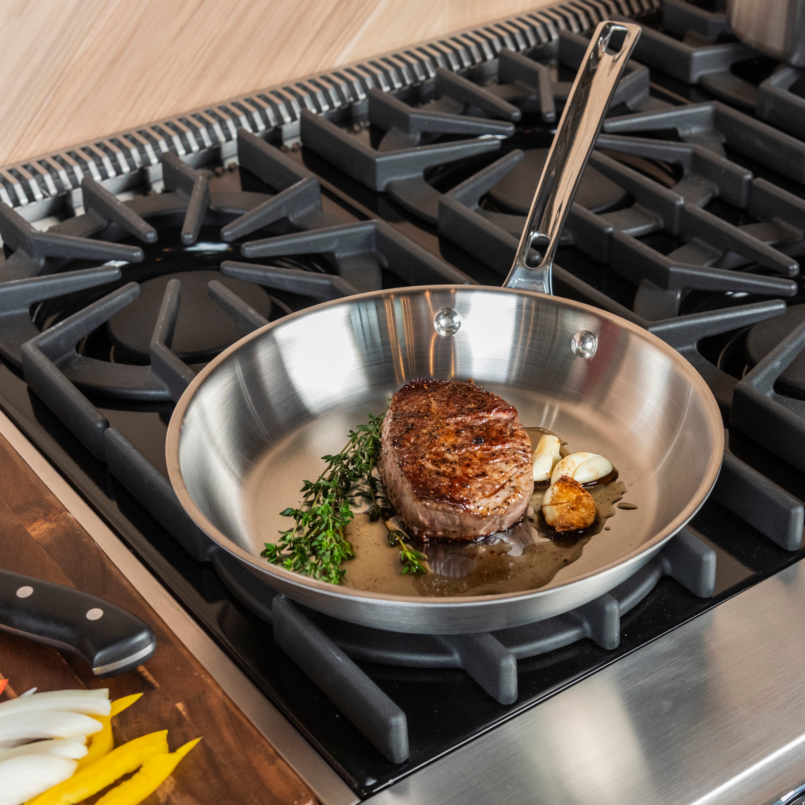 A steak cooking in a stainless steel pan with garlic and herbs on a gas stove, next to a cutting board with sliced vegetables and a knife.
