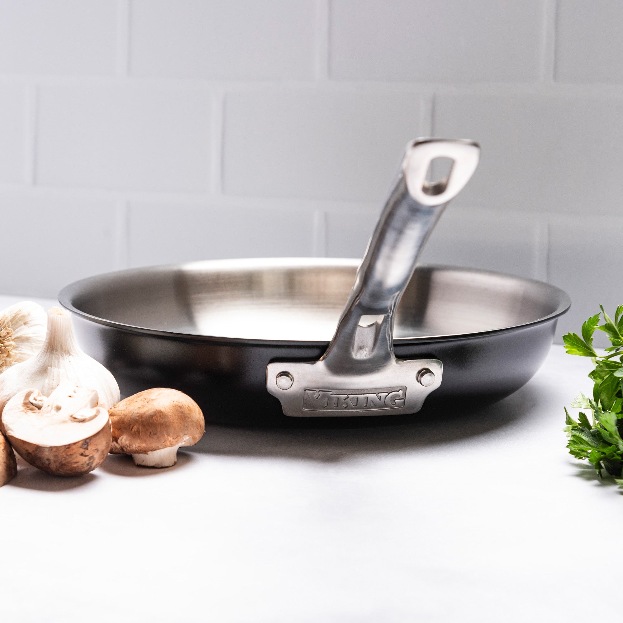 The Viking 12pc 3-Ply Cast Black Exterior Stainless Steel Cookware Set by Viking Culinary Products sits on a white countertop with mushrooms, garlic, and fresh parsley, against a white tiled wall.