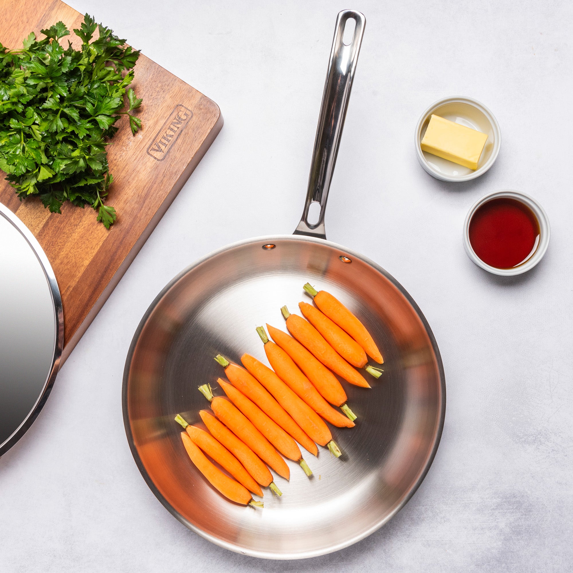 A Viking Culinary Products 12pc 3-Ply Cast Black Exterior Stainless Steel frying pan, filled with peeled baby carrots, rests on a white surface beside bowls of butter and sauce and a cutting board with chopped parsley.