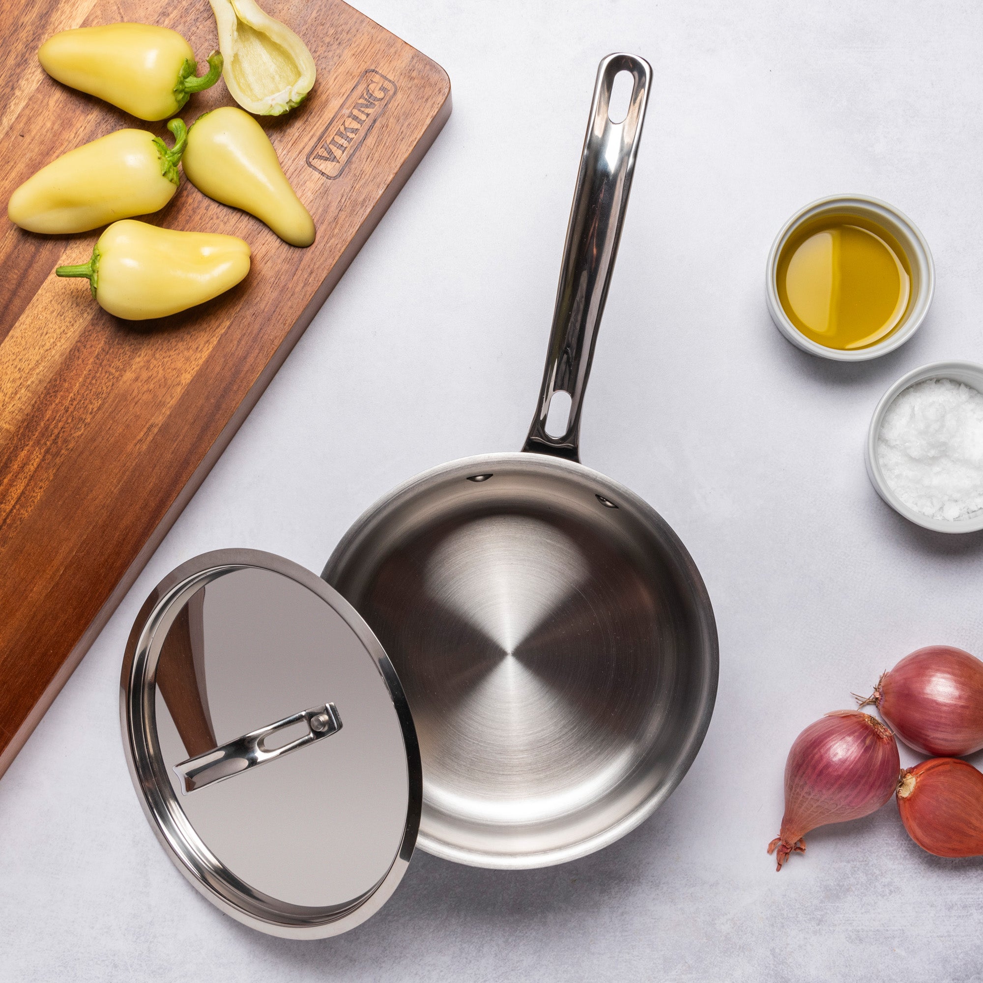 A Viking Culinary Products 12pc 3-Ply Cast Black Exterior Stainless Steel saucepan with lid sits on a light surface, surrounded by shallots, olive oil, salt, a cutting board with yellow peppers, and halved garlic. PFAS free.