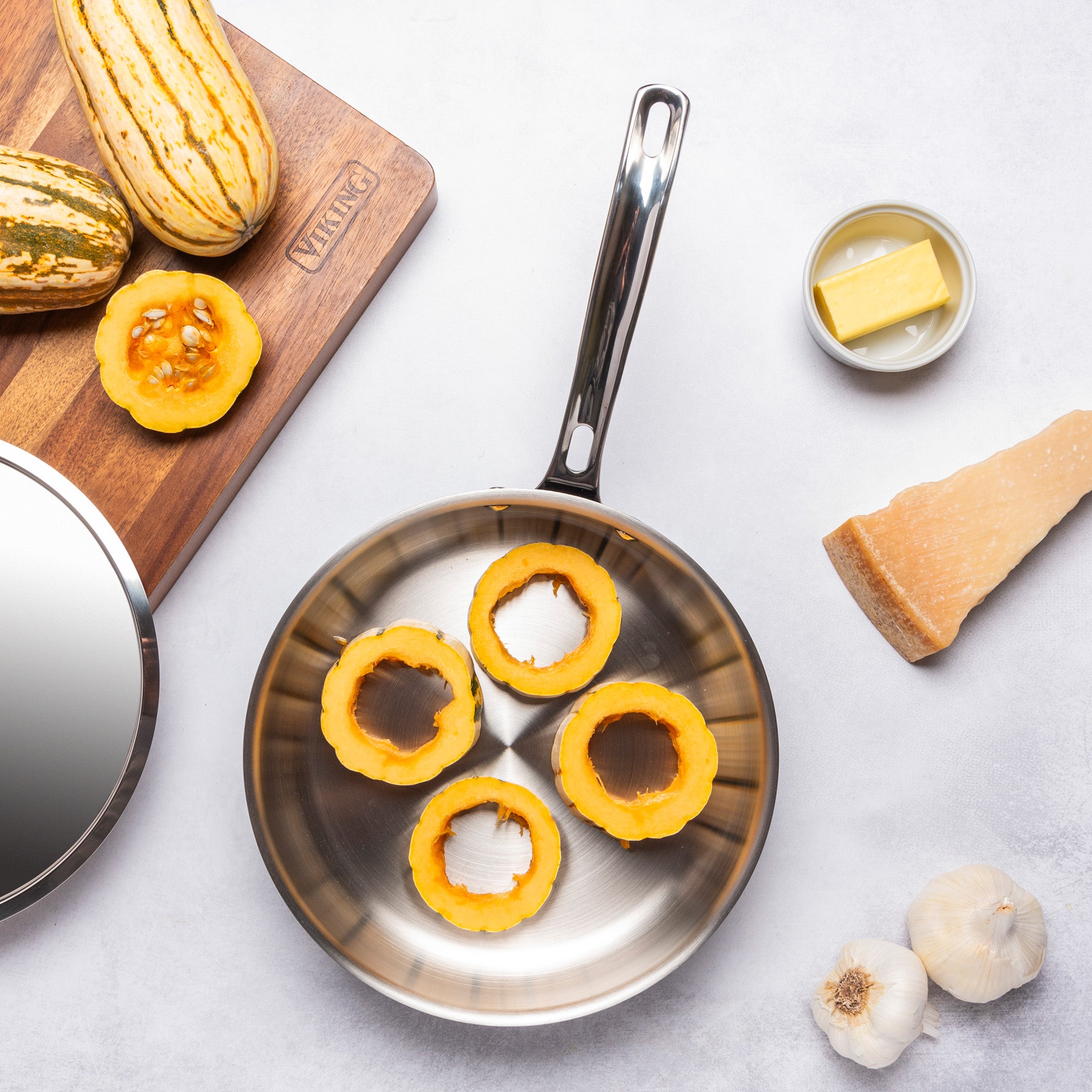 Sliced delicata squash rings in a PFAS-free Viking 12pc 3-Ply Cast Black Exterior Stainless Steel Cookware Set by Viking Culinary Products, shown induction compatible, with whole squash, Parmesan wedge, butter, and garlic on a light background.