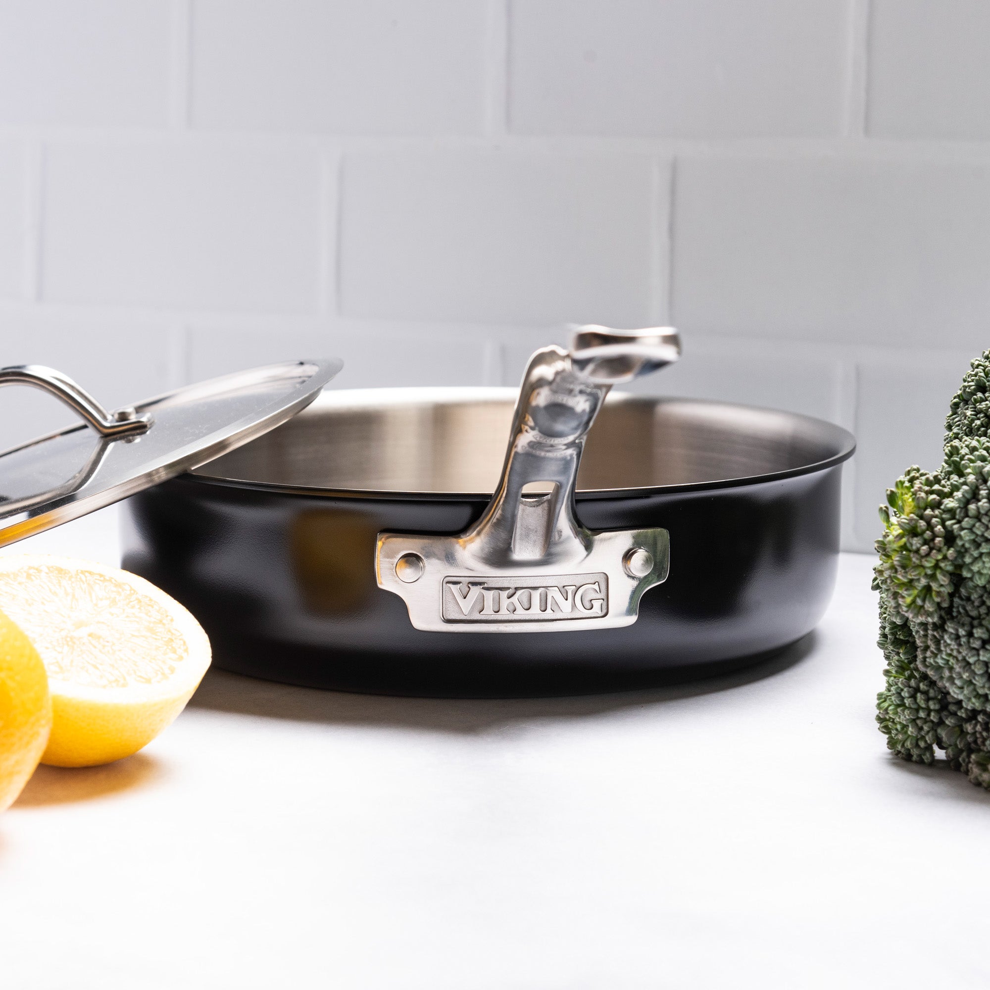 A Viking 12pc 3-Ply Cast Black Exterior Stainless Steel pan with lid from Viking Culinary Products, featuring an aluminum core, sits on a white surface next to broccoli and halved lemons against a white tiled background.