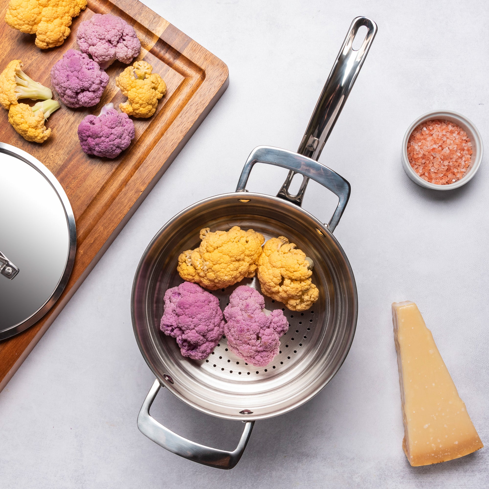 A PFAS-free Viking Culinary Products 12pc 3-Ply Cast Black Exterior Stainless Steel Cookware Set steamer pot with aluminum core holds purple and yellow cauliflower. Nearby, a wooden board displays more florets, cheese wedge, and pink Himalayan salt. Induction compatible.
