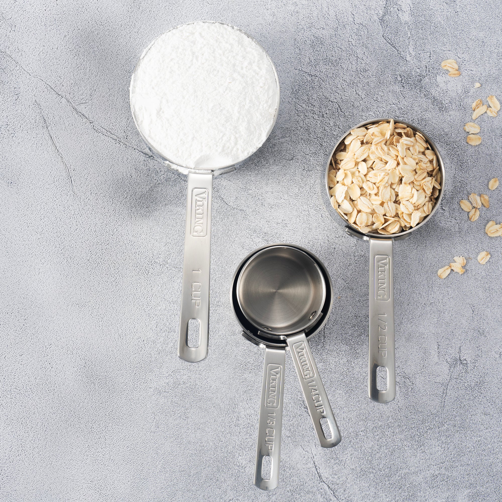The Viking Stainless Steel Measuring Cup Set by Viking Culinary Products, featuring cups labeled 1 cup, 1/2 cup, 1/3 cup, and 1/4 cup, rests on a gray surface. The largest cups hold flour and oats; the smaller ones are empty with oats scattered nearby.