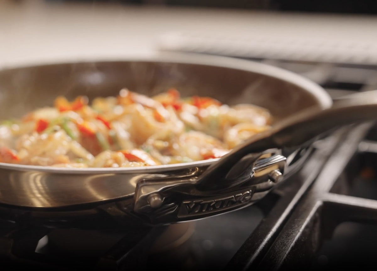 A close-up of a stainless steel skillet with a Viking logo on a stovetop, filled with sautéed food including vegetables and shrimp, with steam rising from the pan.