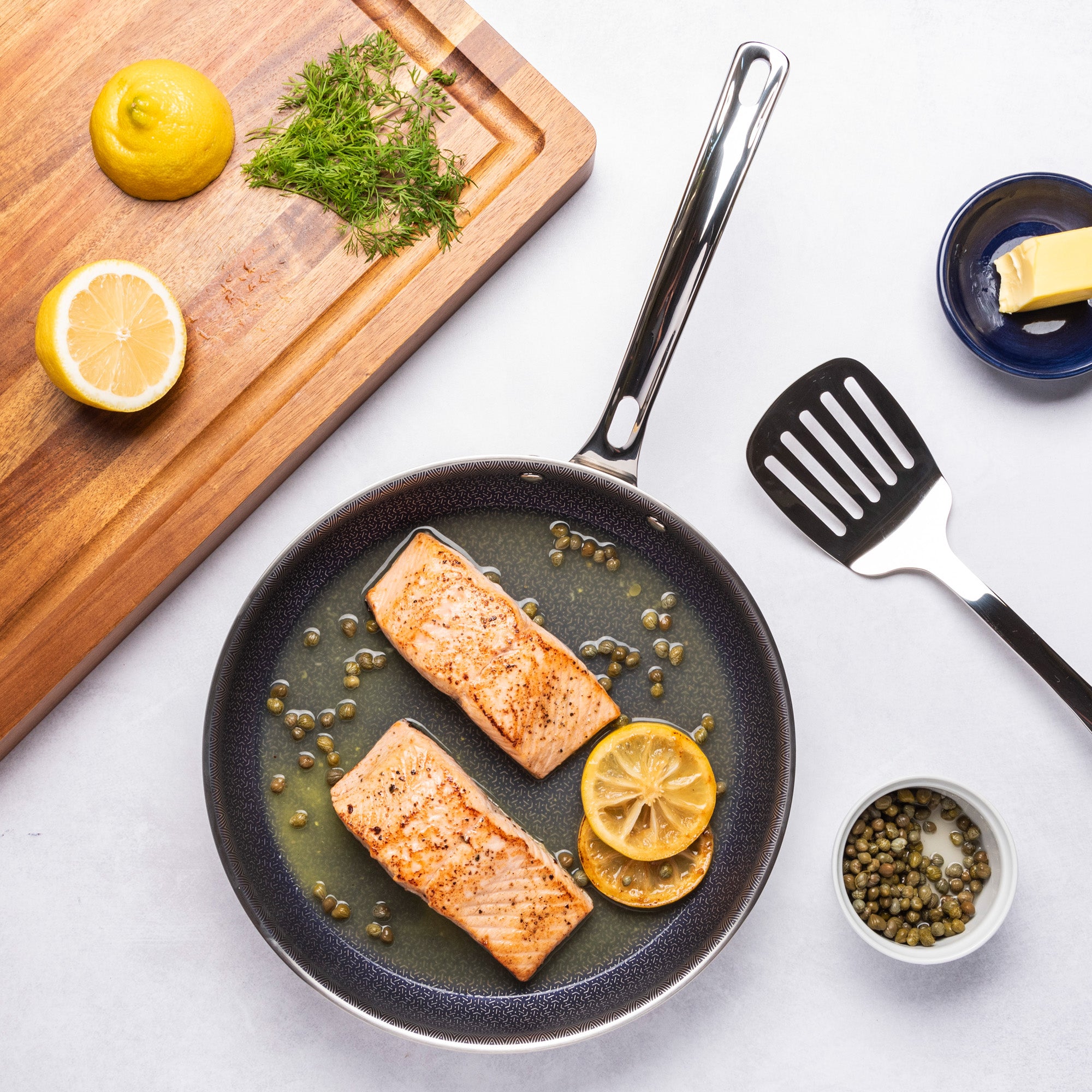 The Viking Culinary Products 3-Ply Hybrid Plus 10-inch Nonstick Fry Pan holds two cooked salmon fillets, lemon slices, and capers. Nearby are a spatula, butter on a plate, extra capers, halved lemon, and fresh dill on a cutting board.