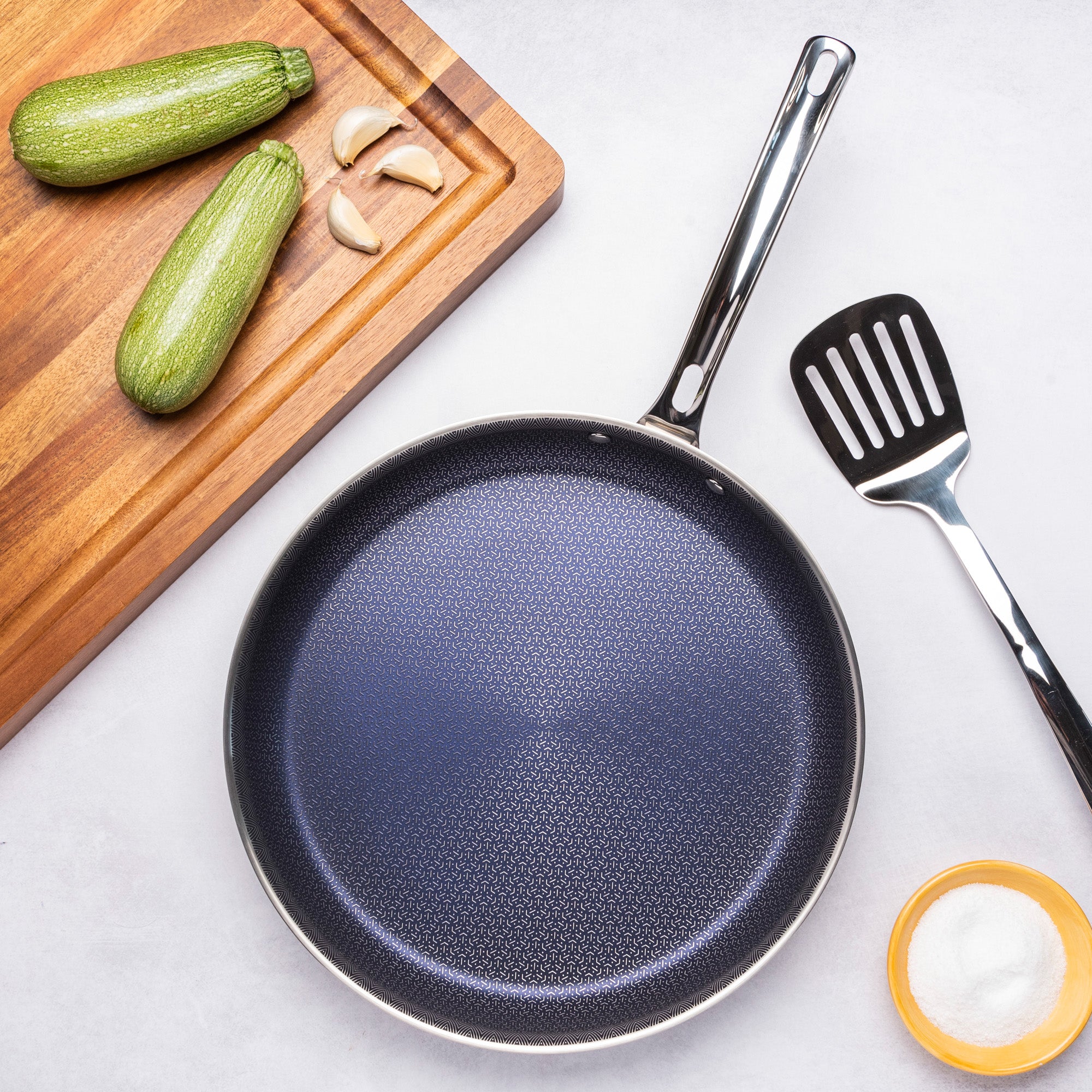 The Viking Culinary Products 3-Ply Hybrid Plus 12-inch Nonstick Fry Pan sits on a light surface with a black spatula, a small bowl of salt, a wooden cutting board holding two zucchinis, and three garlic cloves nearby.