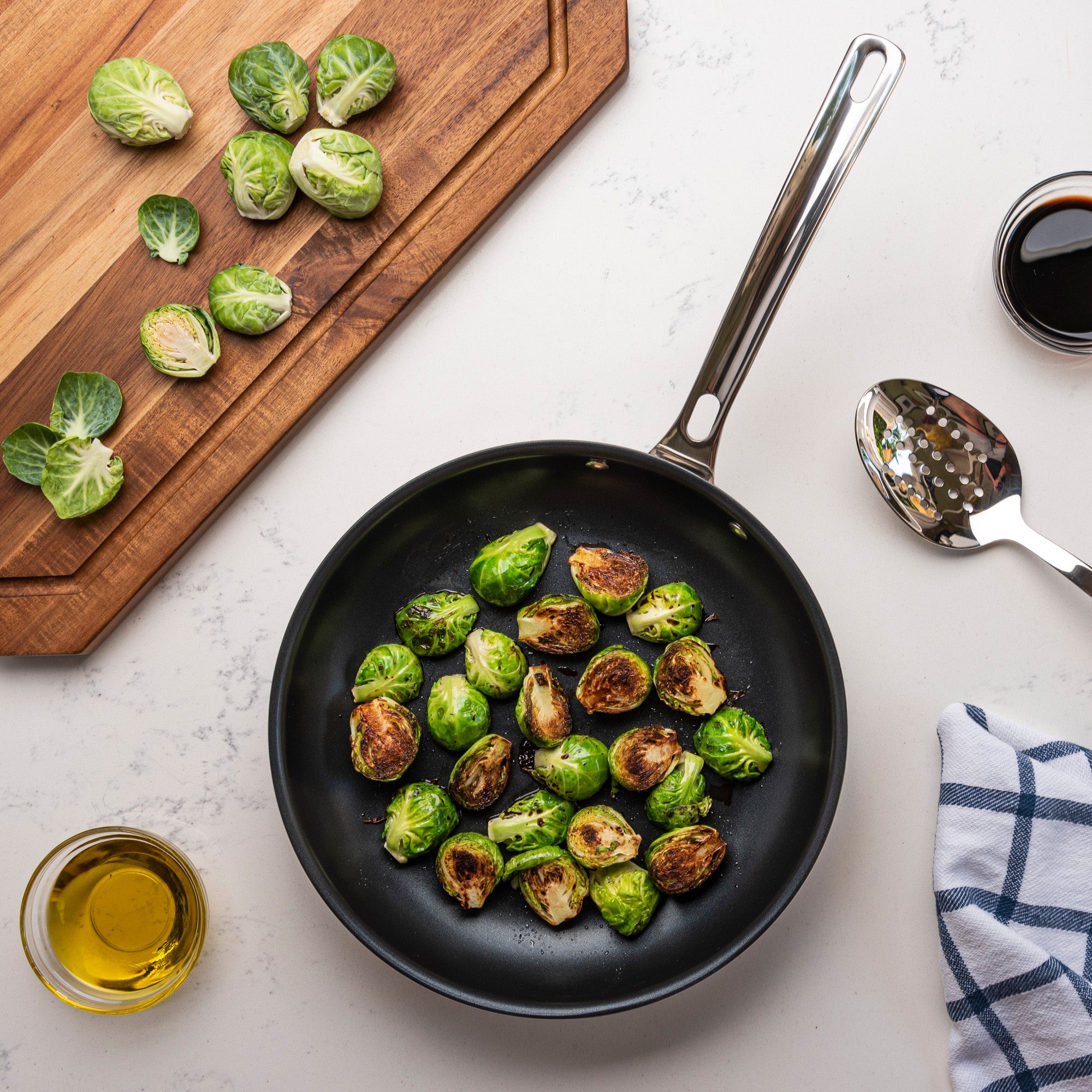 A Viking 3-Ply Stainless Steel Nonstick Fry Pan from Viking Culinary Products with roasted Brussels sprouts rests on a white countertop, beside a wooden cutting board with halved sprouts, a spoon, olive oil, sauce dish, and blue-checkered towel.