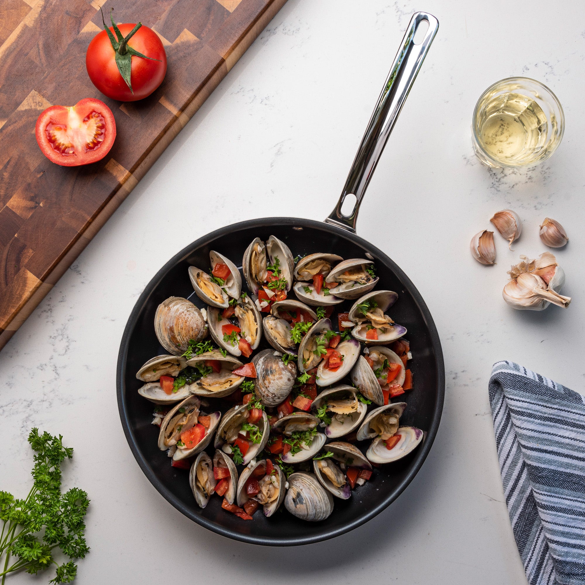 A skillet of clams with tomatoes and herbs rests on a white counter with parsley, garlic, wine, and a towelâ€”prepared in the Viking Culinary Products 3-Ply Stainless Steel 2-Piece Nonstick Fry Pan Set, which is dishwasher safe for convenience.