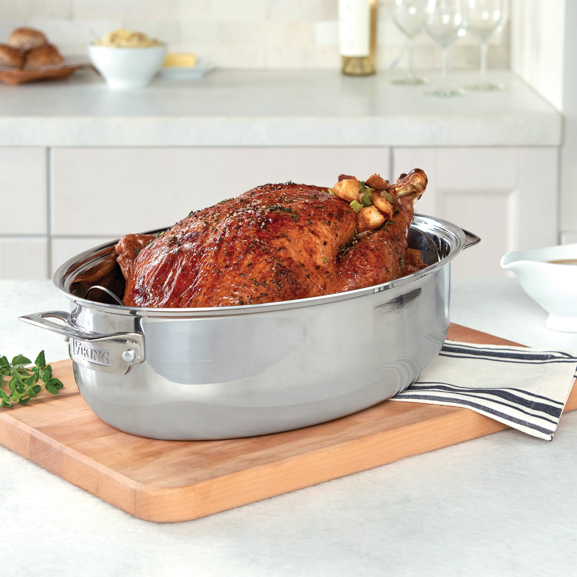 A golden-brown turkey stuffed with herbs rests in the Viking 3-Ply 8.5-Quart 3-in-1 Oval Roaster by Viking Culinary Products, set on a wooden board in a modern kitchen. A striped towel peeks out, accenting the induction-compatible cookwareâ€™s sleek design.