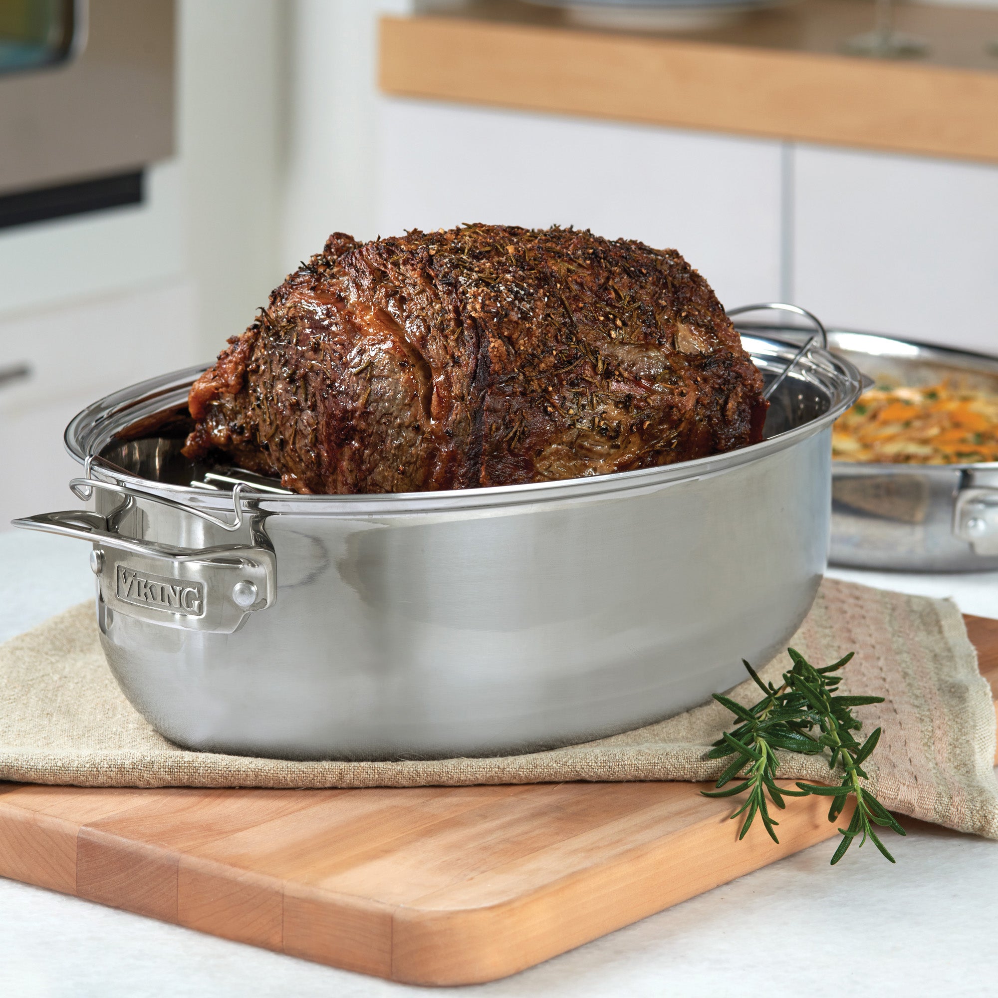 A roasted prime rib rests in the Viking 3-Ply 8.5-Quart 3-in-1 Oval Roaster with Rack by Viking Culinary Products on the counter, surrounded by fresh rosemary and a casserole dish, showcasing induction-compatible cookware.