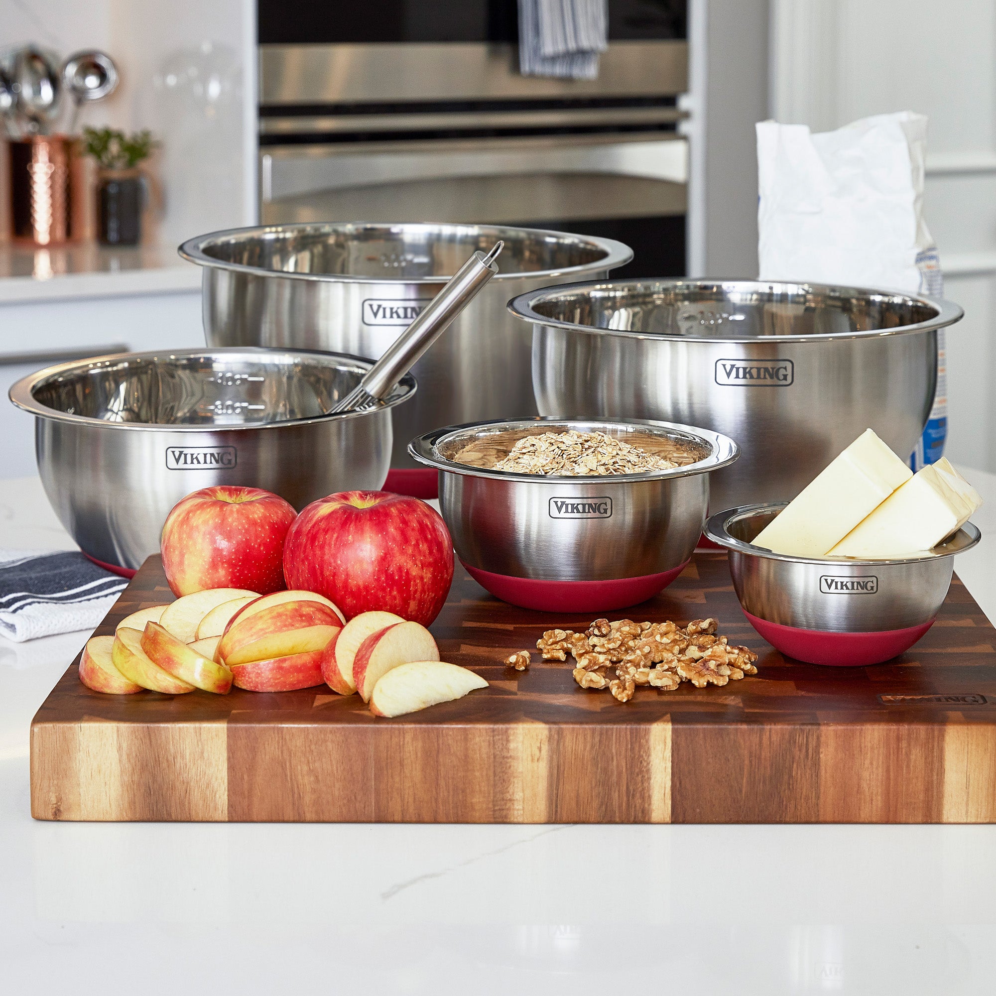 The Viking Culinary Products 10-Piece Stainless Steel Mixing Bowl Set with Red Lids is displayed in a modern kitchen, holding apples, apple slices, walnuts, butter, and flour on a wooden board. A whisk sits in one bowl for easy prep and storage.
