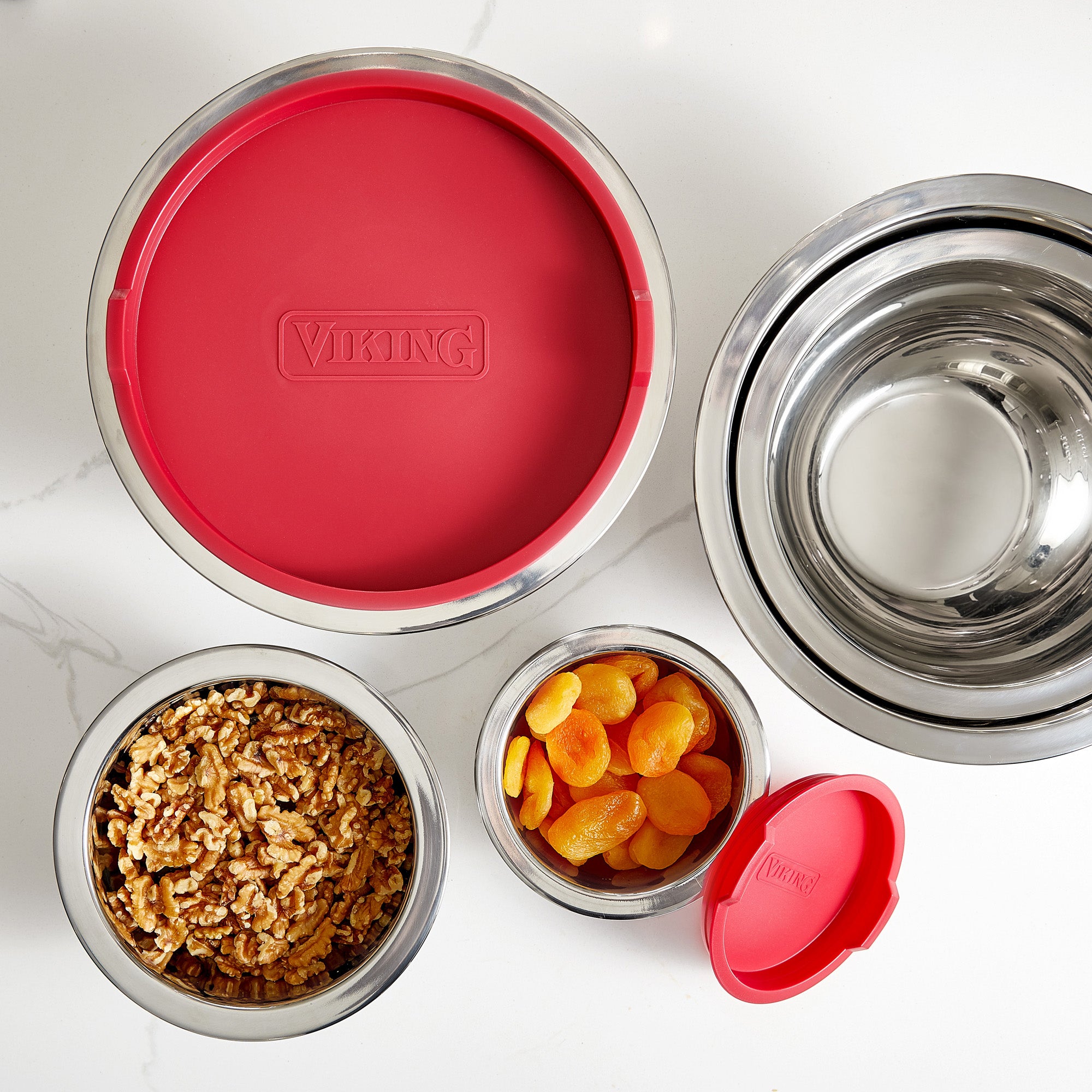 The Viking 10-Piece Stainless Steel Mixing Bowl Set with Red Lids by Viking Culinary Products is shown on a white surface; two bowls have airtight lids, one labeled VIKING, and the bowls contain walnuts and dried apricots.