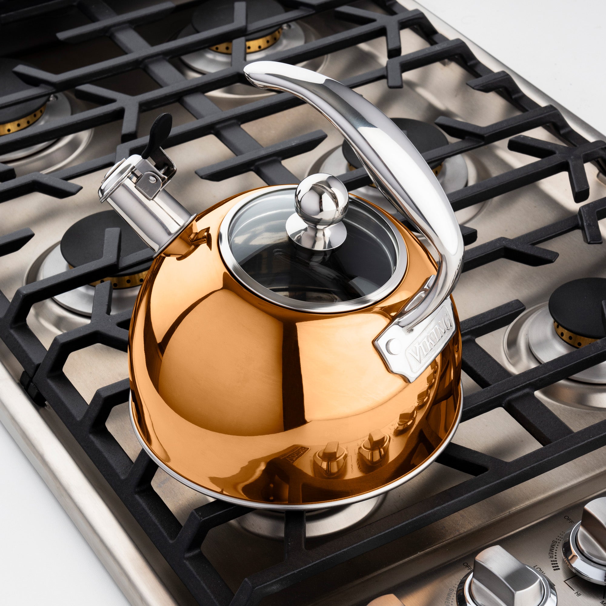 The Viking 2.6-Quart Rose Gold Stainless Steel Whistling Kettle by Viking Culinary Products sits on a stove, its silver handle gleaming, ready to boil water and alert you with a whistle when it’s done.