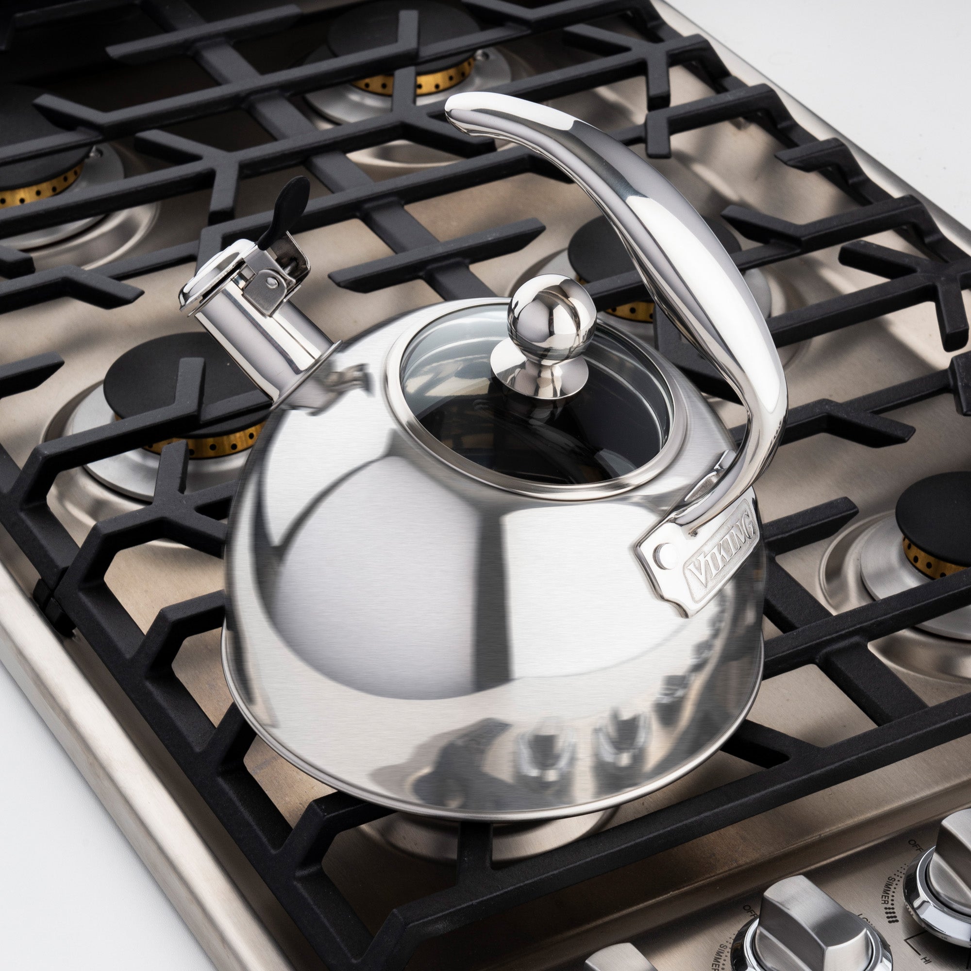 A Viking Culinary Products 2.6-Quart Satin Finish Stainless Steel Whistling Kettle with 3-Ply Base sits on a modern gas stove with four burners and black grates, handle raised and spout cover open in a clean, contemporary kitchen.