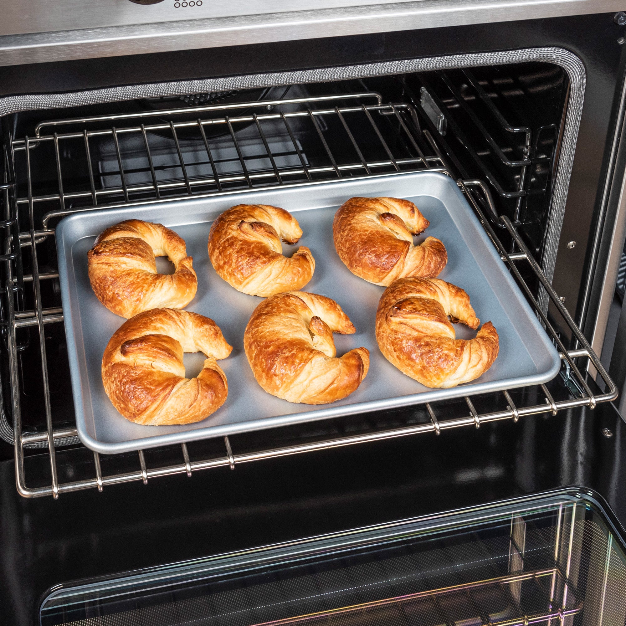 Six golden-brown croissants bake on a Viking 15-Inch Aluminized Nonstick Baking Sheet by Viking Culinary Products, placed on the middle oven rack with the oven door open.