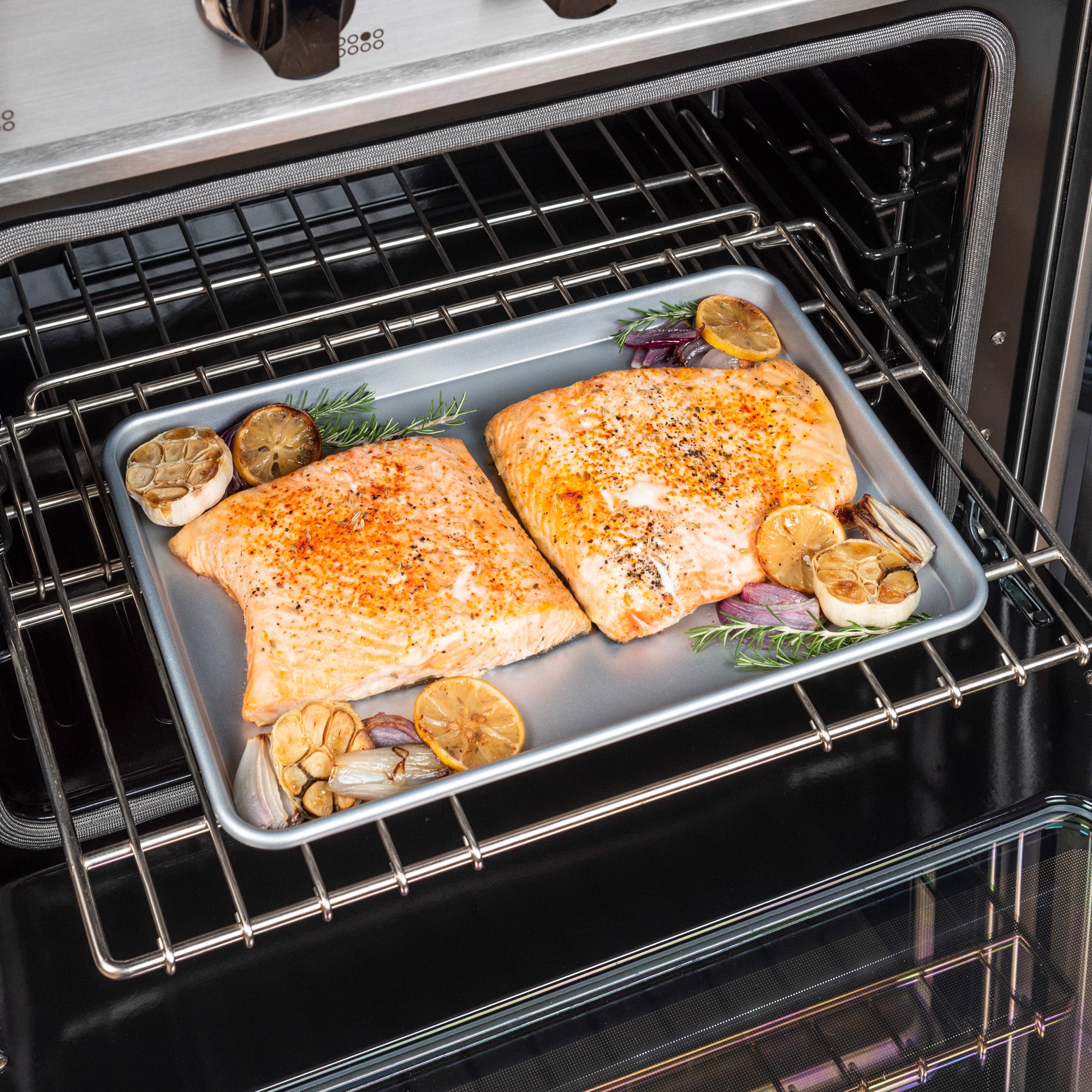 Two seasoned salmon fillets with lemon, garlic, rosemary, and onions bake on the Viking Culinary Products 2-Piece Nonstick Aluminized Steel Baking Sheet Set, arranged on an oven rack.