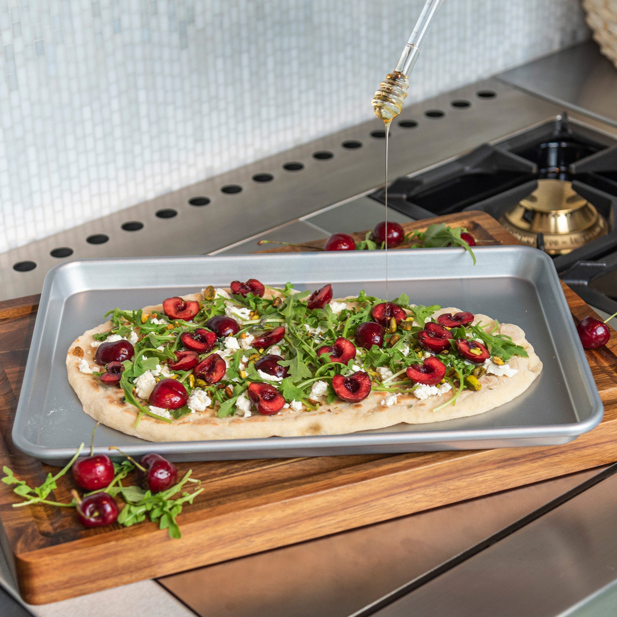 A flatbread with cherries, arugula, cheese, and pistachios is served on the Viking 15-Inch Aluminized Nonstick Baking Sheet by Viking Culinary Products. Honey is drizzled on top, with extra cherries and greens for garnish.