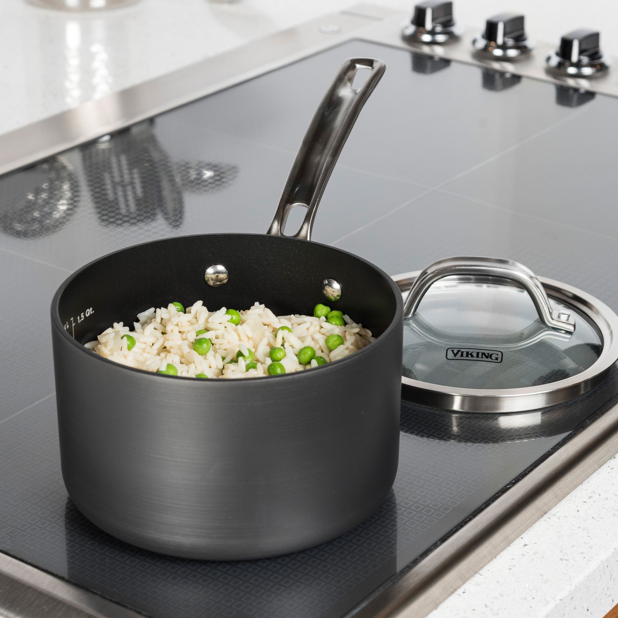 A Viking Culinary Products Hard Anodized Nonstick 2-Quart Sauce Pan with rice and green peas sits on a glass stovetop beside its clear glass lid. Stove dials and another pot are also visible in the background.