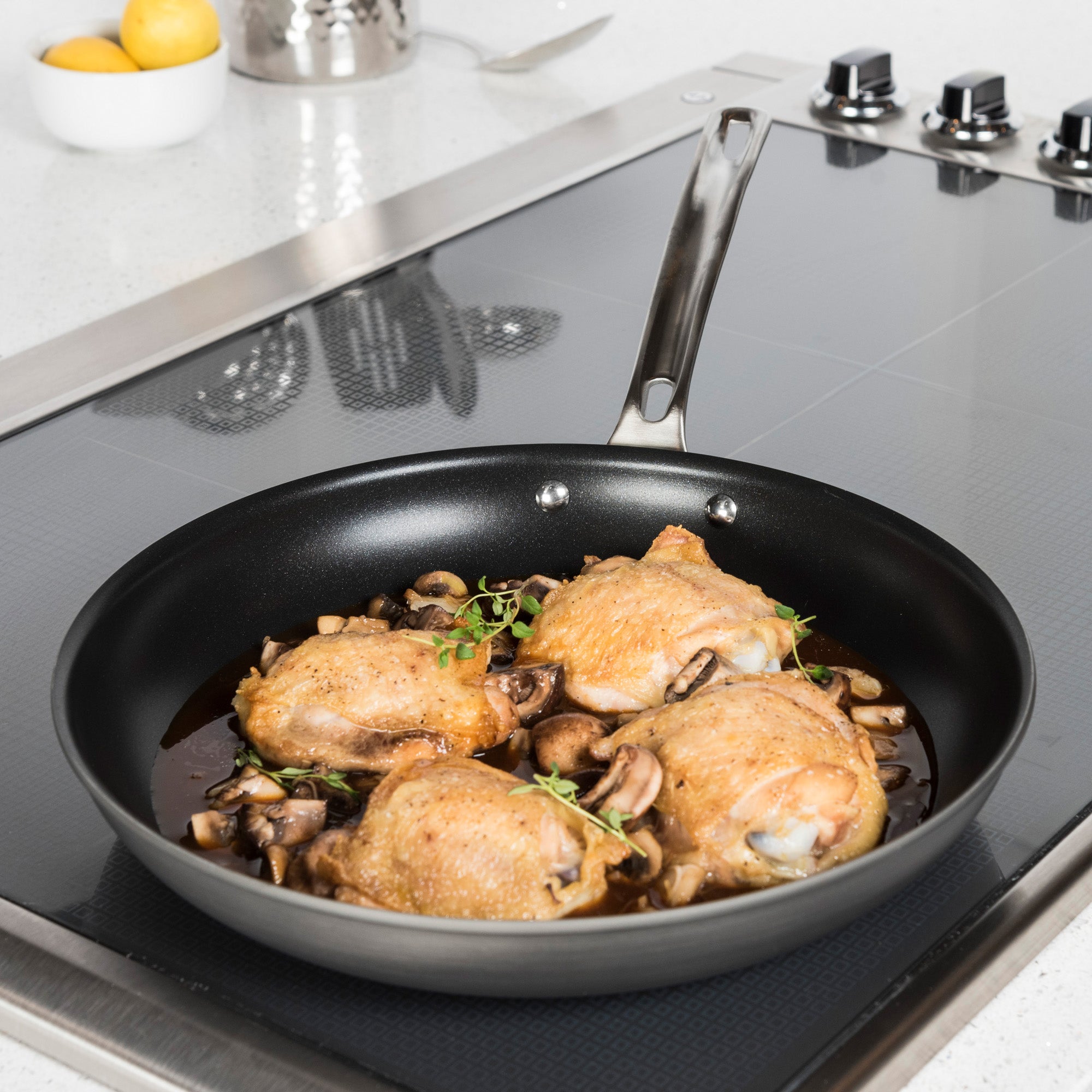 A Viking Culinary Products Hard Anodized Nonstick skillet from the 10-Piece Cookware Set with Glass Lids holds cooked chicken thighs and mushrooms on a modern stovetop, while a Dutch oven rests on a white countertop nearby.