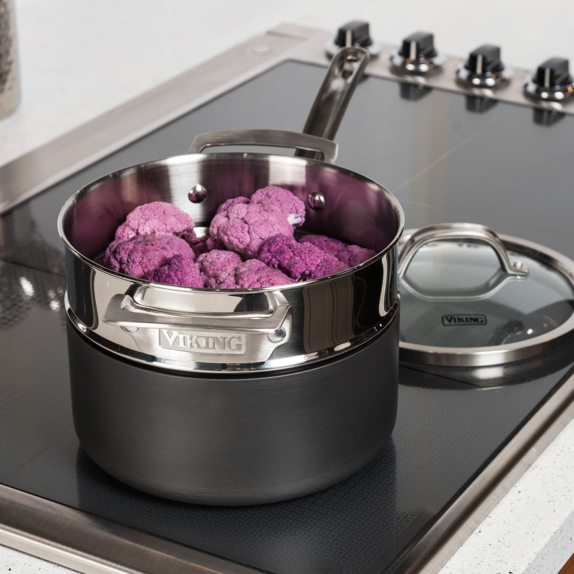 A Viking Stainless Steel Universal Steamer Insert and Colander by Viking Culinary Products sits atop a modern electric stovetop, resting in a pot of purple cauliflower with metal handles and a glass lidâ€”ideal for versatile steaming.