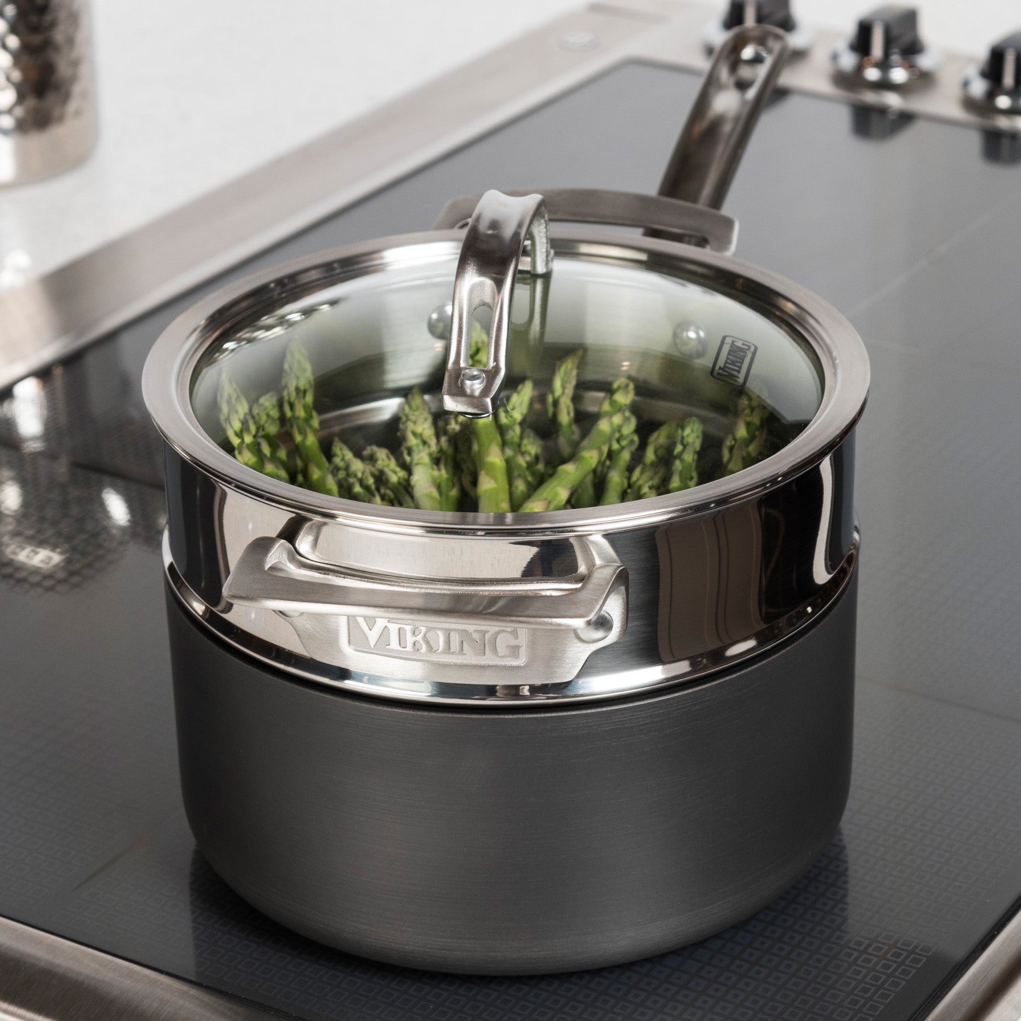 The Viking Stainless Steel Universal Steamer Insert and Colander from Viking Culinary Products steams fresh green asparagus spears on the stove, its metal handles and glass lid visible.